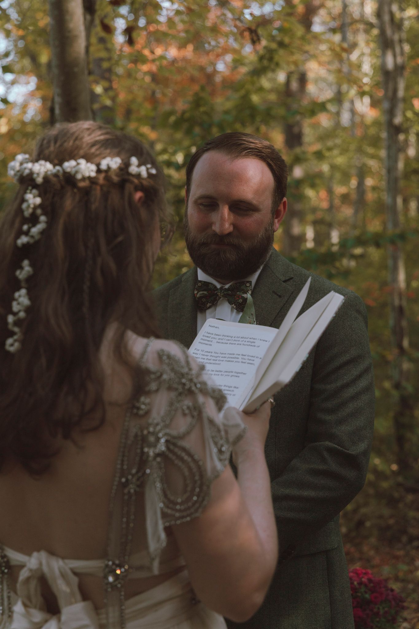 A couple shares a tender moment reading their vows together in an autumn forest setting with fall foliage in the background.
