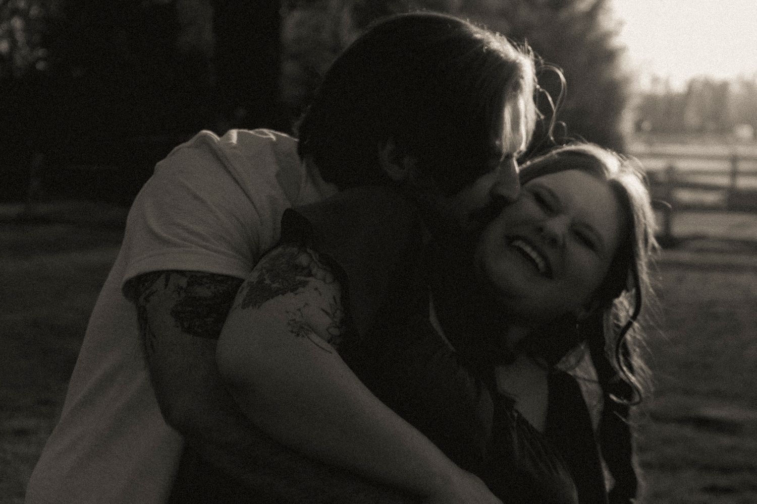 Black and white photo of a couple embracing outdoors, man kissing woman's cheek as she laughs joyfully.