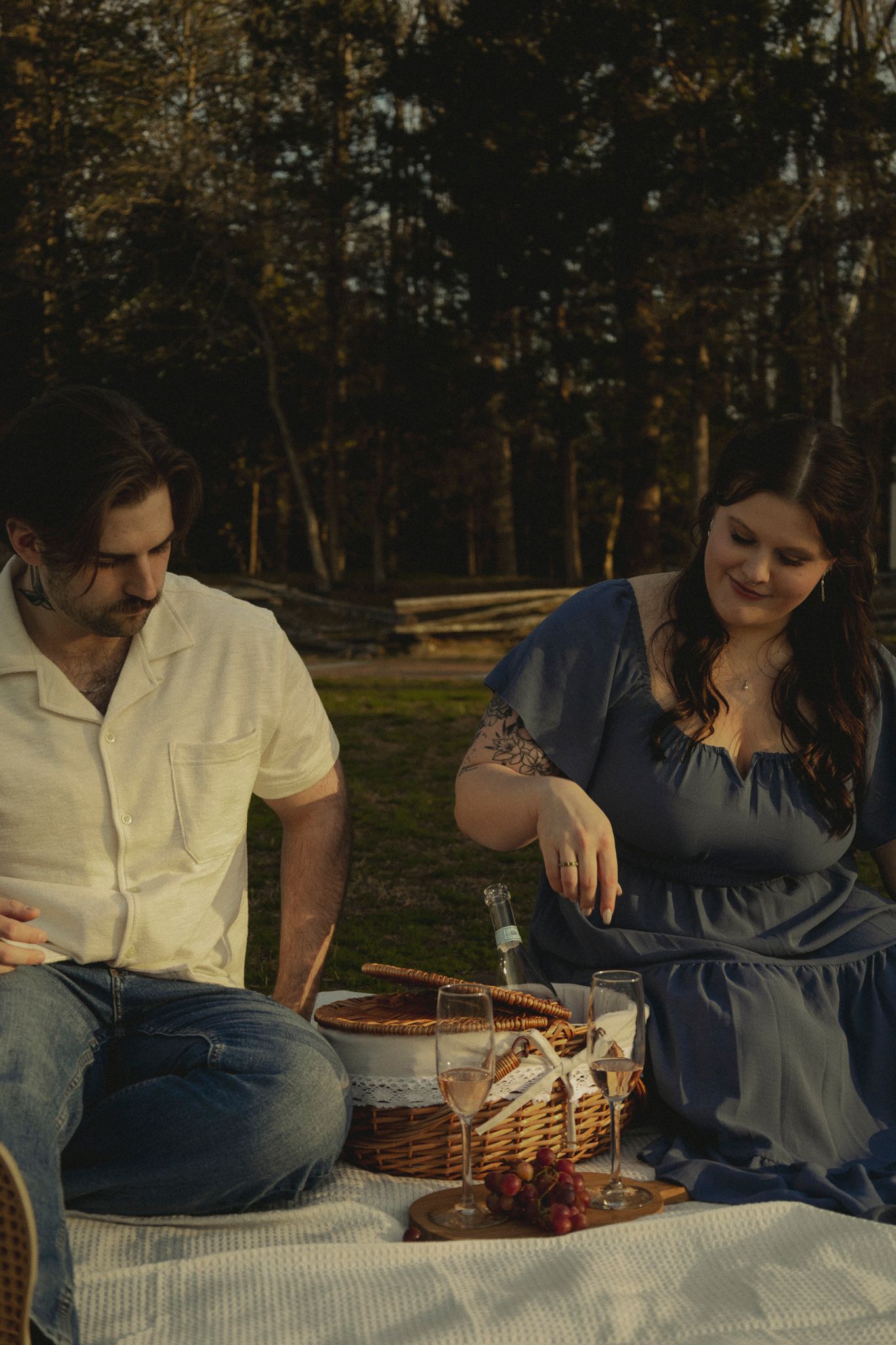 Couple enjoying a romantic outdoor picnic on a blanket with wine and food in a sunlit forest clearing.