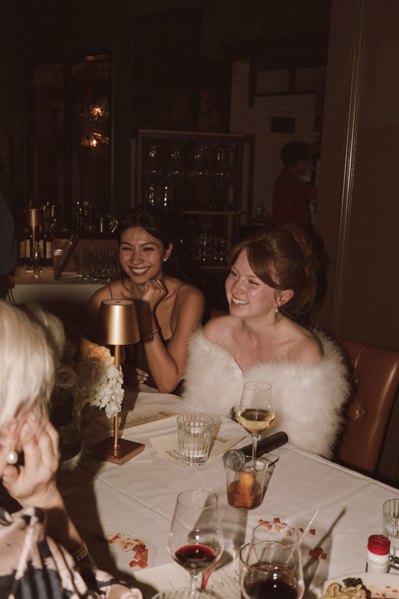 Bride in white gown with feather wrap laughs joyfully with dark-haired guest at reception table.
