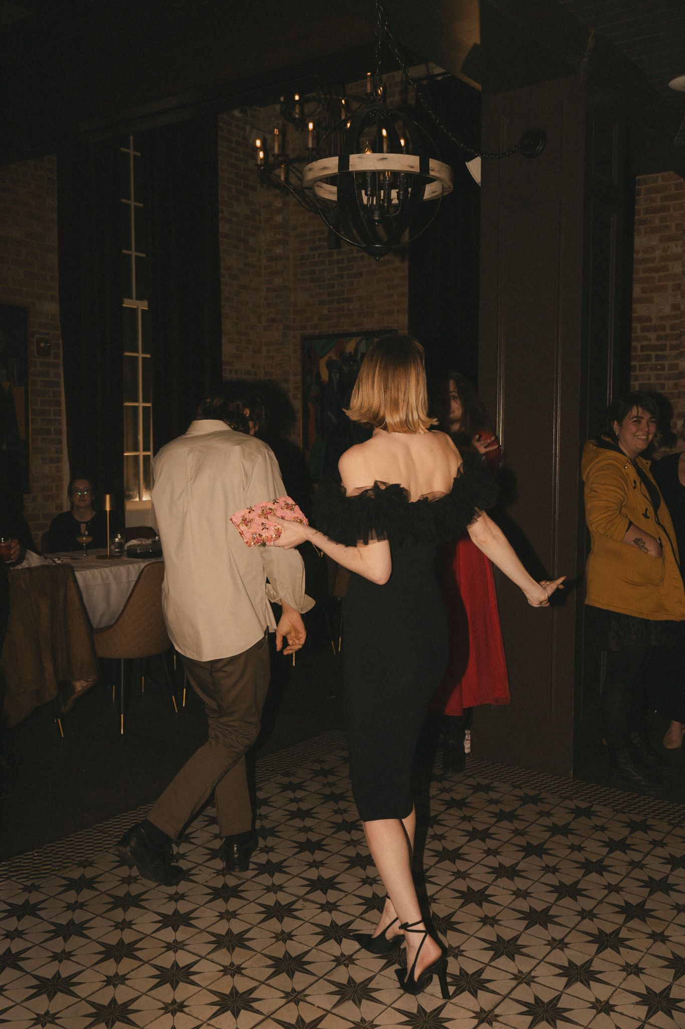 Guests dancing at wedding reception under chandelier in rustic brick-walled venue.