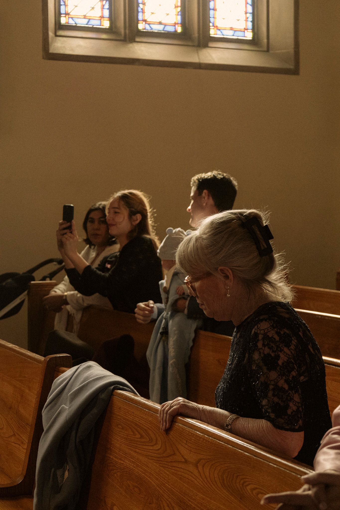 Wedding guests in church pews, with a young woman taking photos on her smartphone during the ceremony.