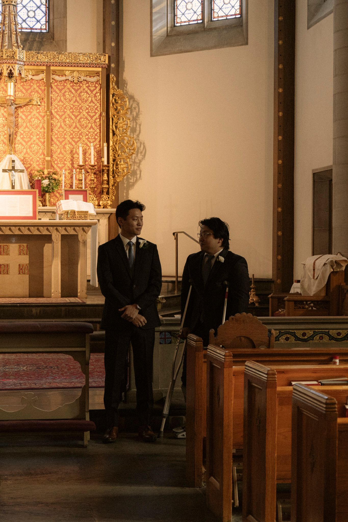 Two groomsmen in dark suits stand near the altar of an ornate church before a wedding ceremony.