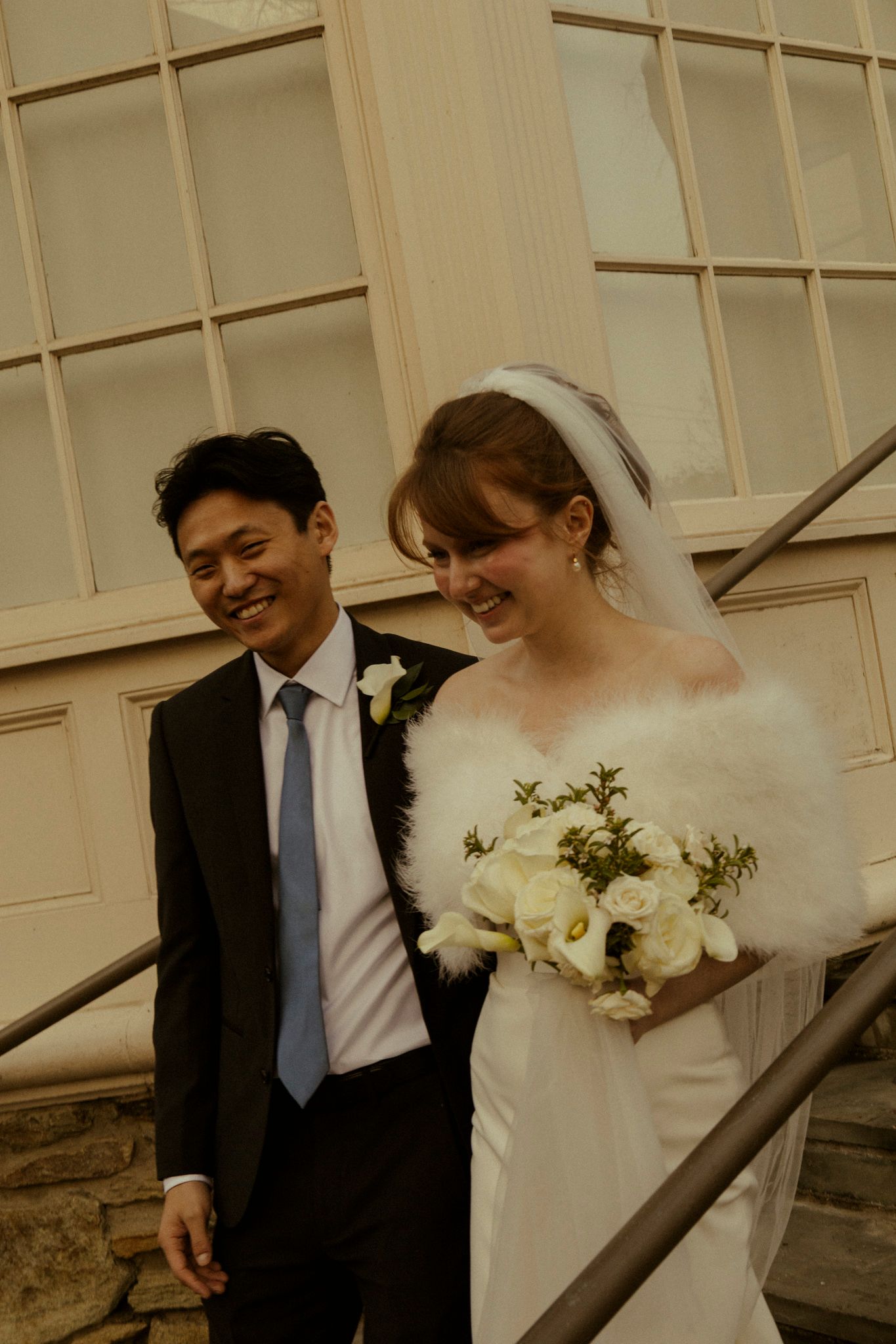 Happy bride in white dress, fur wrap, and veil holds bouquet beside smiling groom in black suit on stone steps.