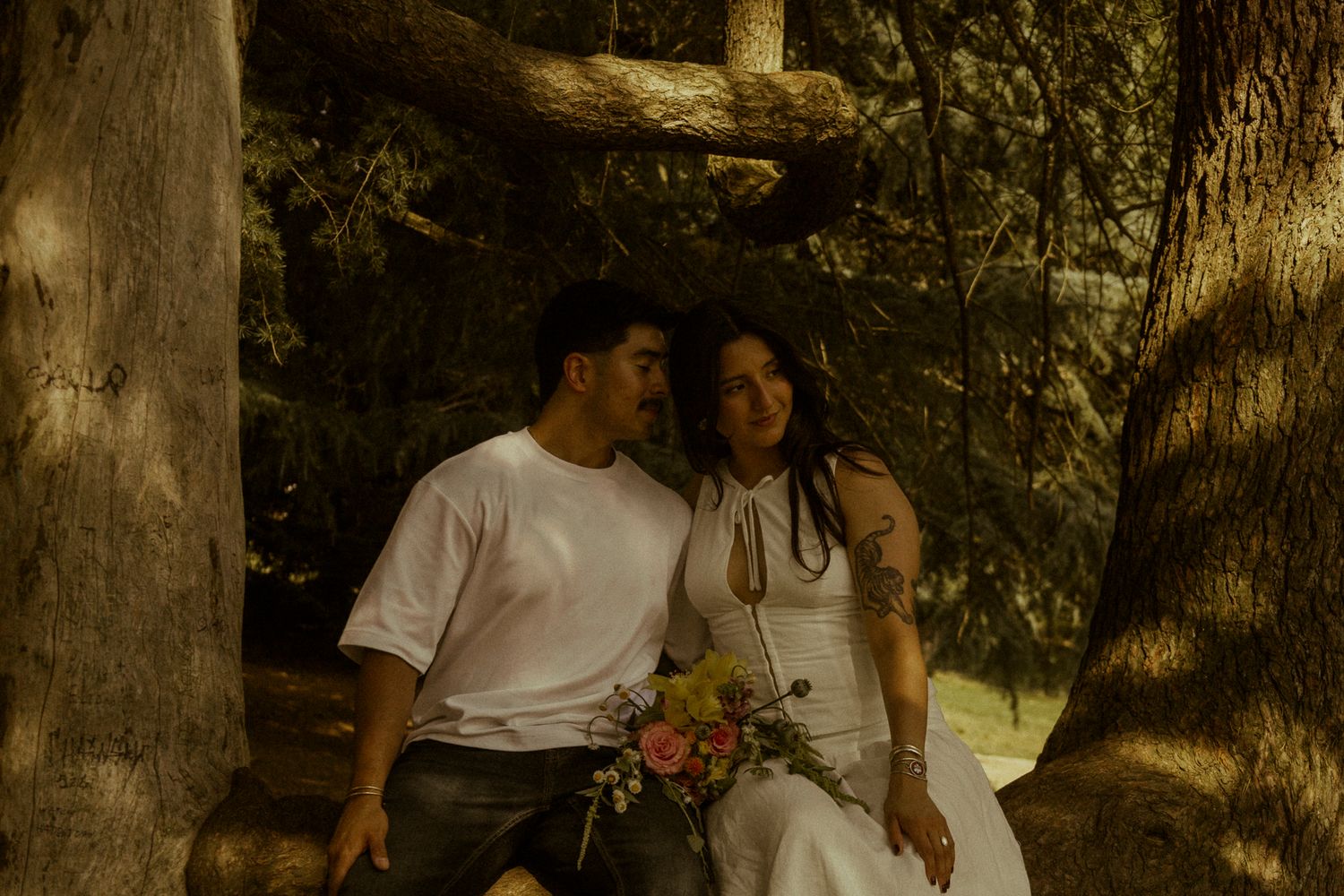 Couple sitting together among trees, woman in white dress holding colorful bouquet, man in white shirt, romantic outdoor setting.