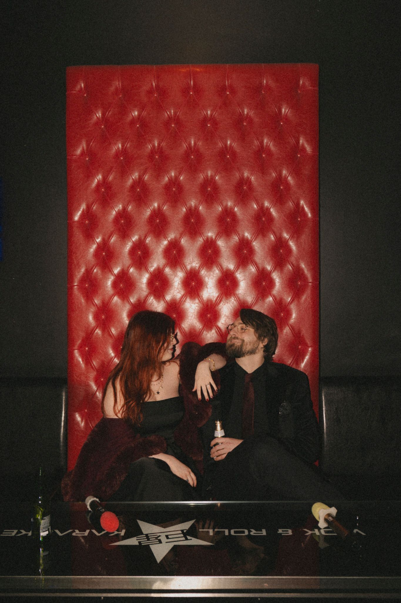 A couple sits intimately on a red tufted leather booth in a dimly lit venue.