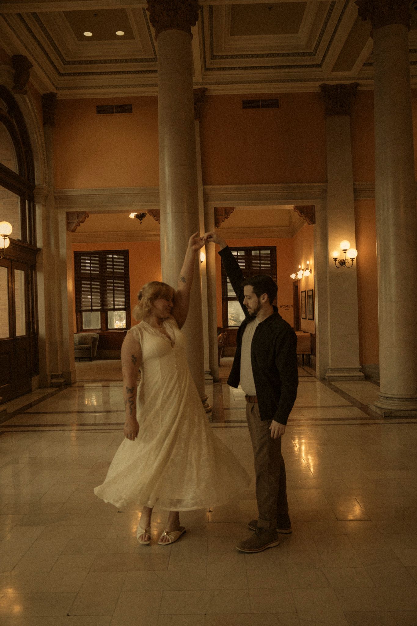 A couple dances together in an elegant ballroom with ornate columns and warm lighting creating a romantic atmosphere.