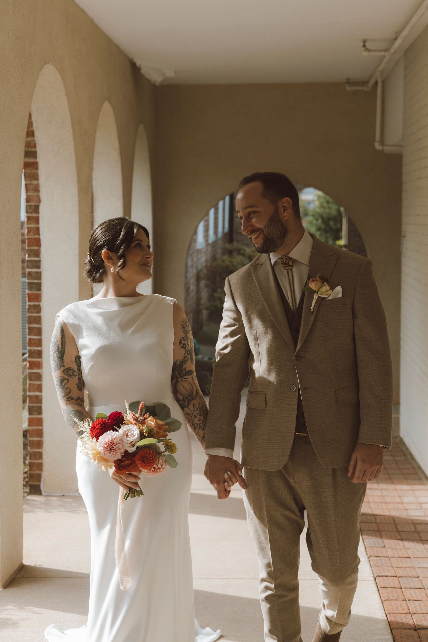 A couple in wedding attire walks together through an elegant columned hallway with brick accents.