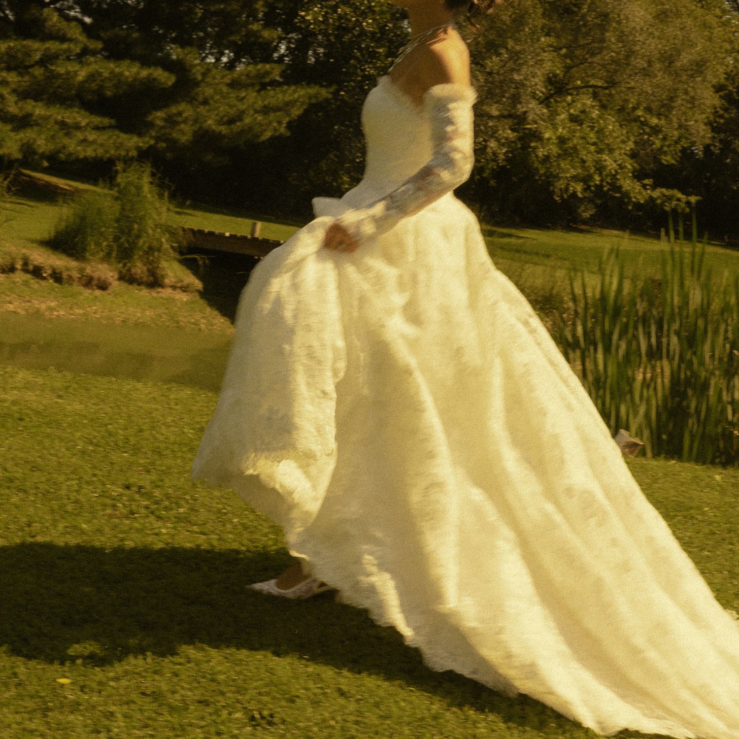 Bride in elegant white lace ball gown with long gloves posing outdoors near a pond in a lush green garden.