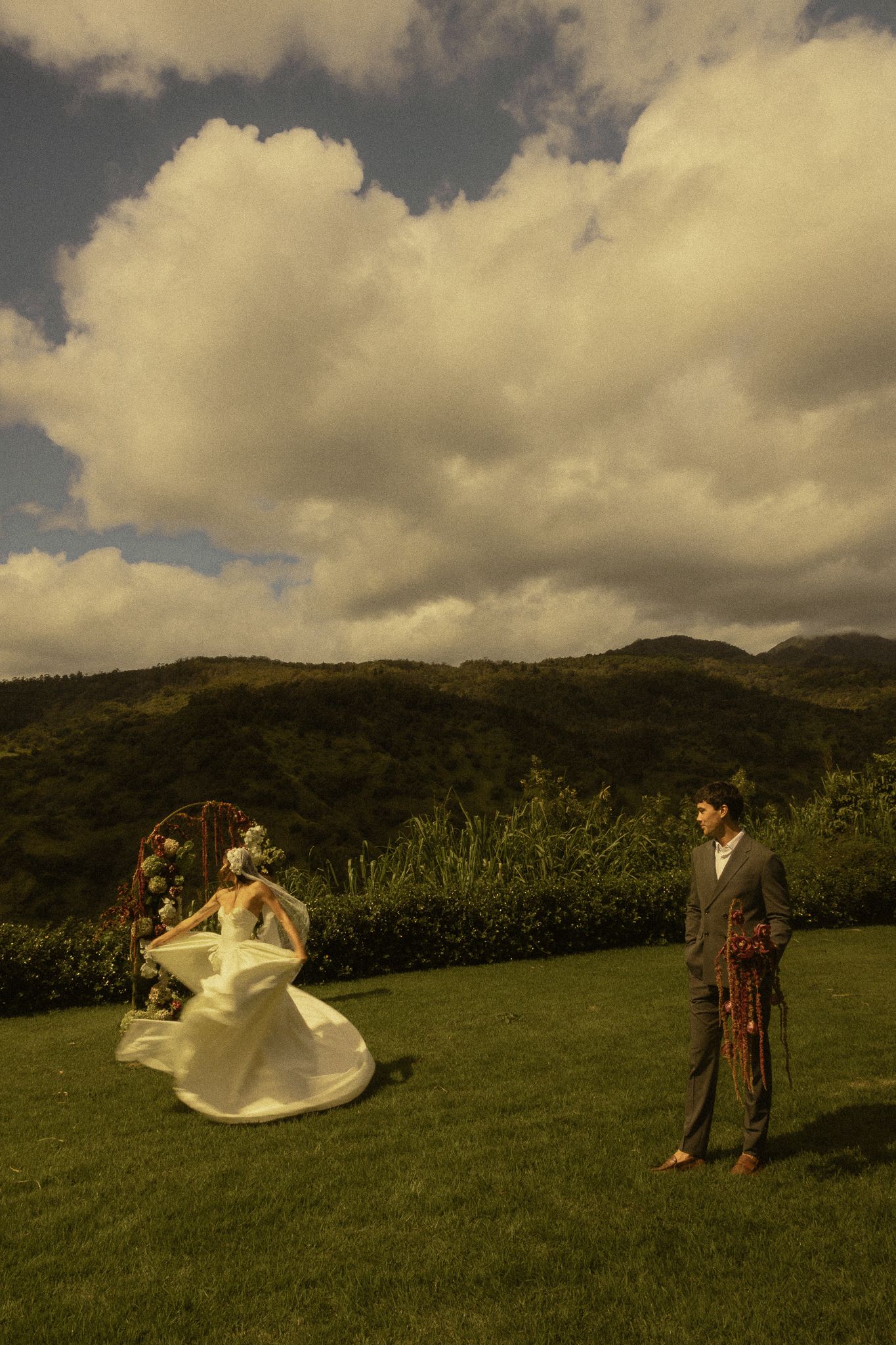 Bride in flowing white dress twirls near floral arch while groom stands on lush green lawn with mountain backdrop.