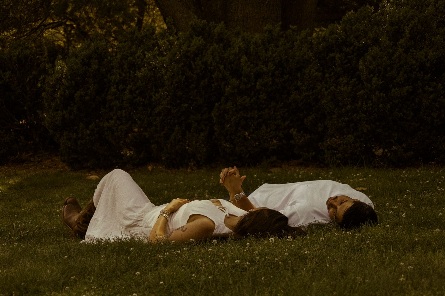Two people in white clothing lying on green grass near dark hedges outdoors.