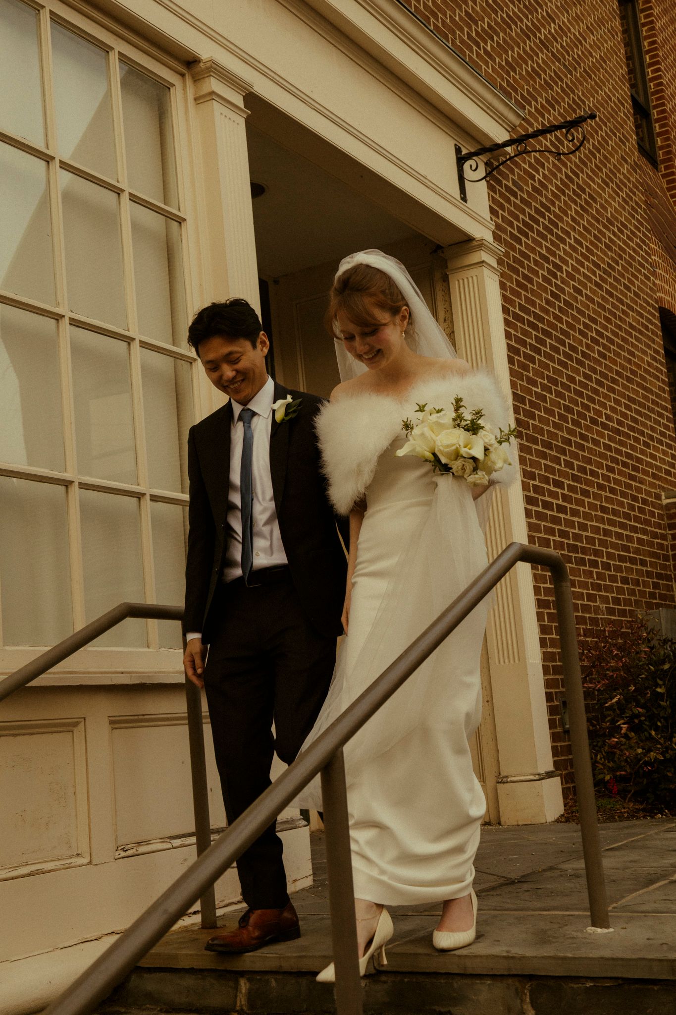 Bride and groom descend church steps, she wears fur wrap and veil, he wears dark suit.