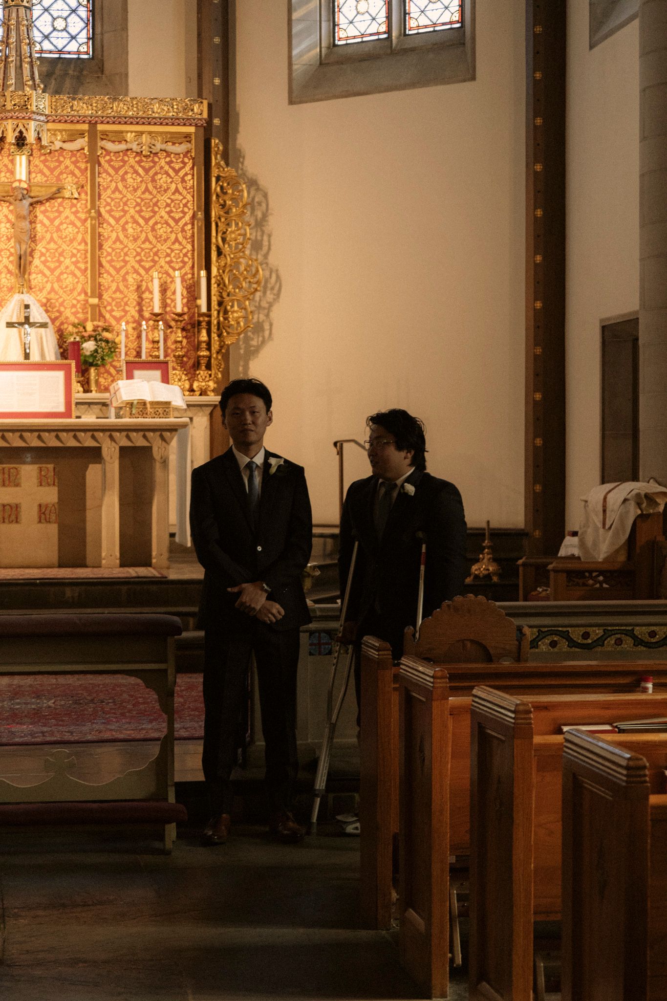 Two men in dark suits stand in a dimly lit church interior with ornate altar and wooden pews.