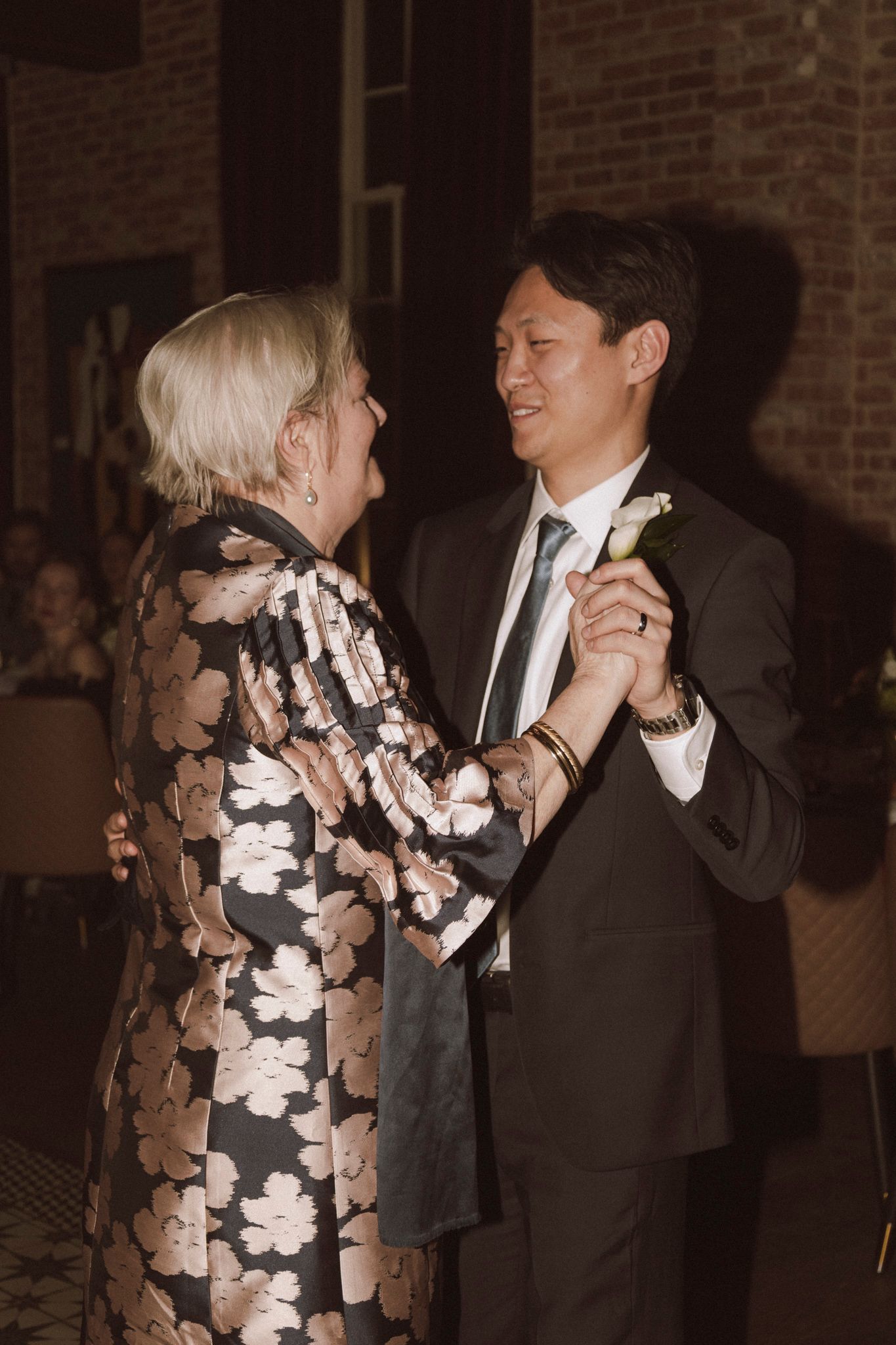 Groom in dark suit dancing with older woman in floral dress during mother-son wedding dance at reception.