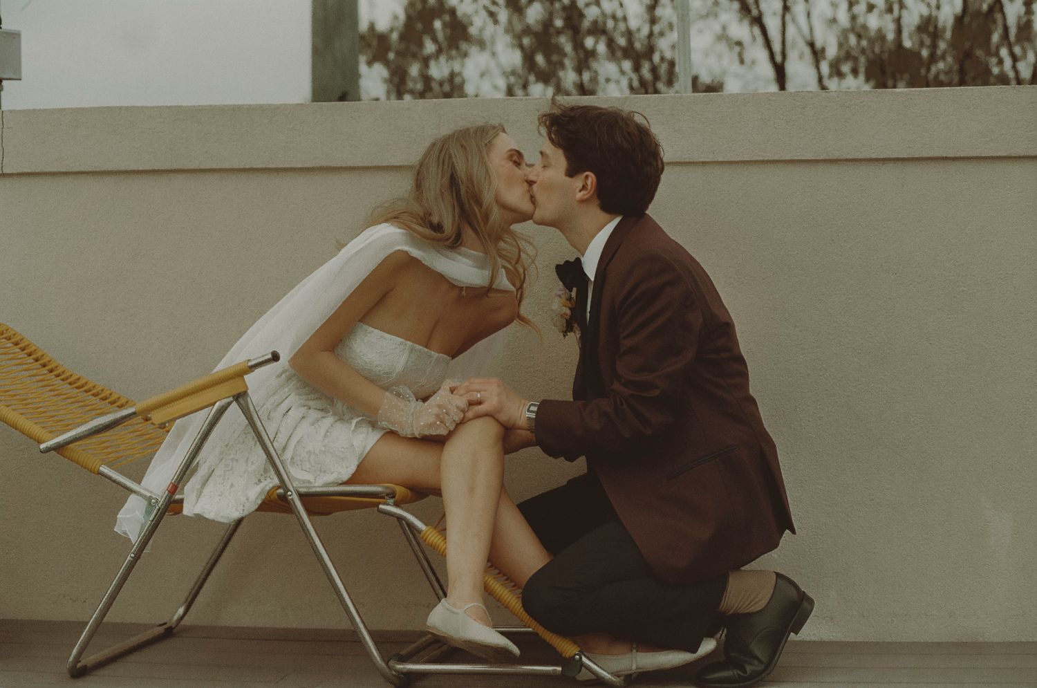 A couple in vintage clothing share an intimate moment on a folding chair against a neutral wall.