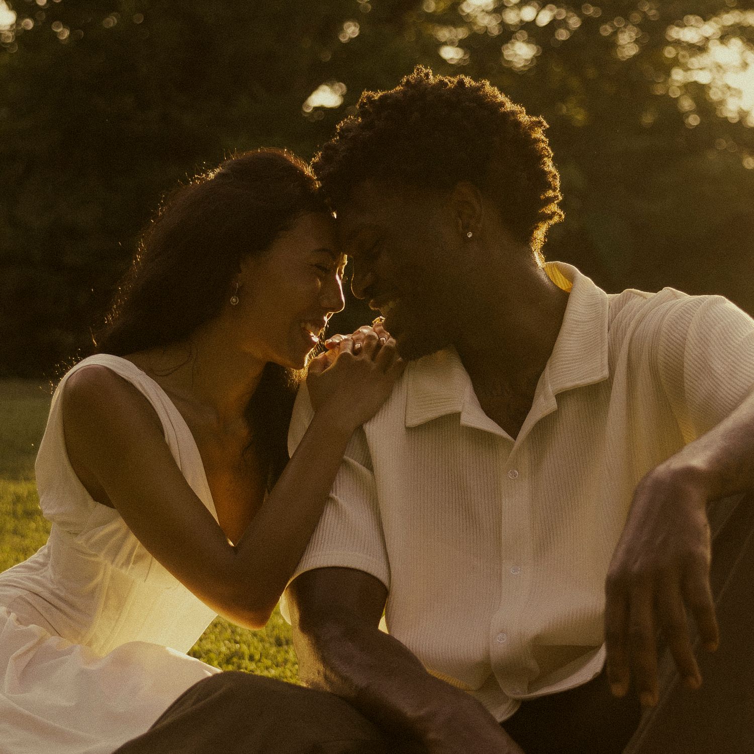 A couple shares a romantic golden hour moment outdoors, leaning close together while sitting on grass.