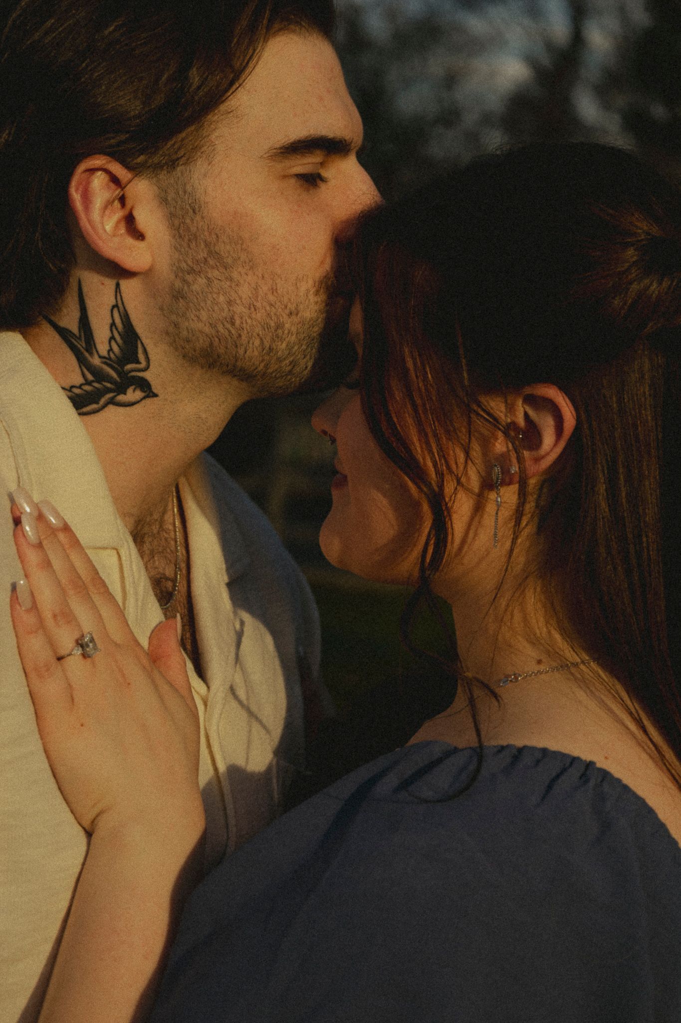 Couple sharing a tender kiss outdoors at golden hour, man with bird neck tattoo embracing woman.