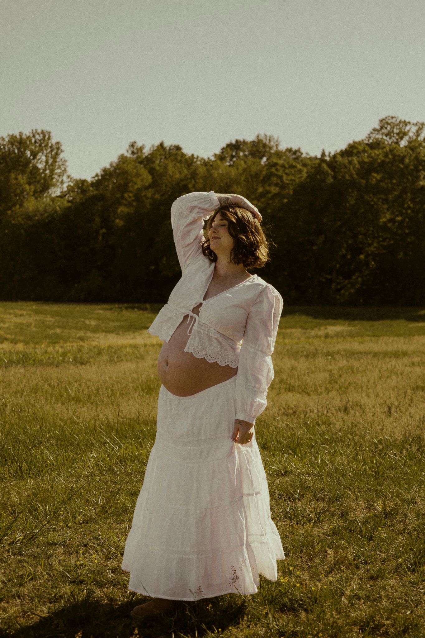 Pregnant woman in white crop top and maxi skirt posing in sunlit green field during golden hour maternity photoshoot.