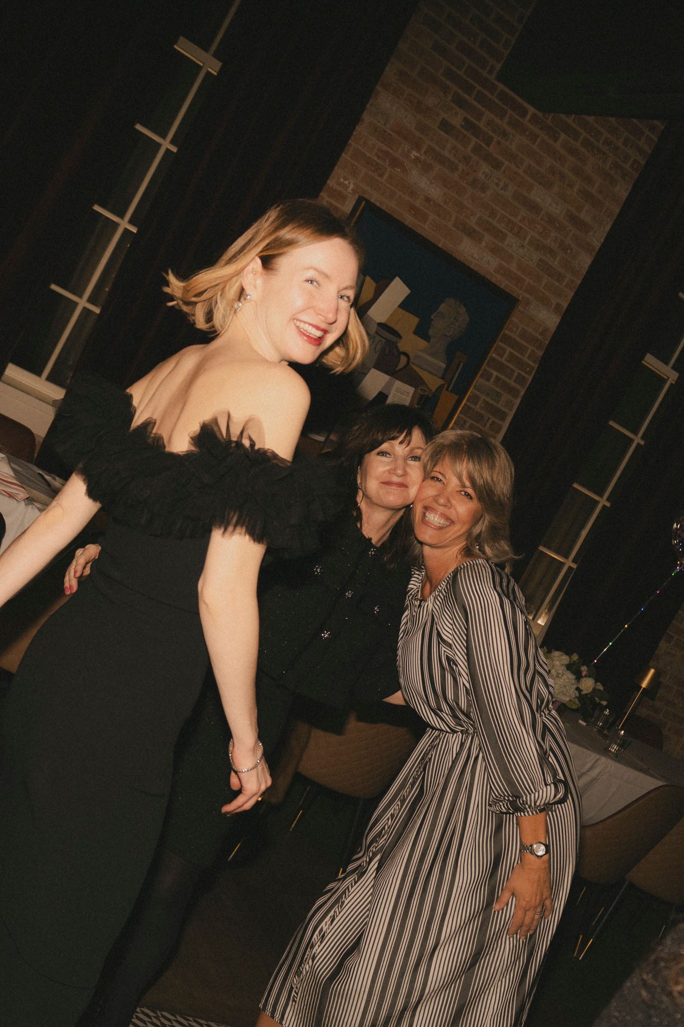 Three women smiling and posing together at an elegant evening event, one wearing a black off-shoulder dress.