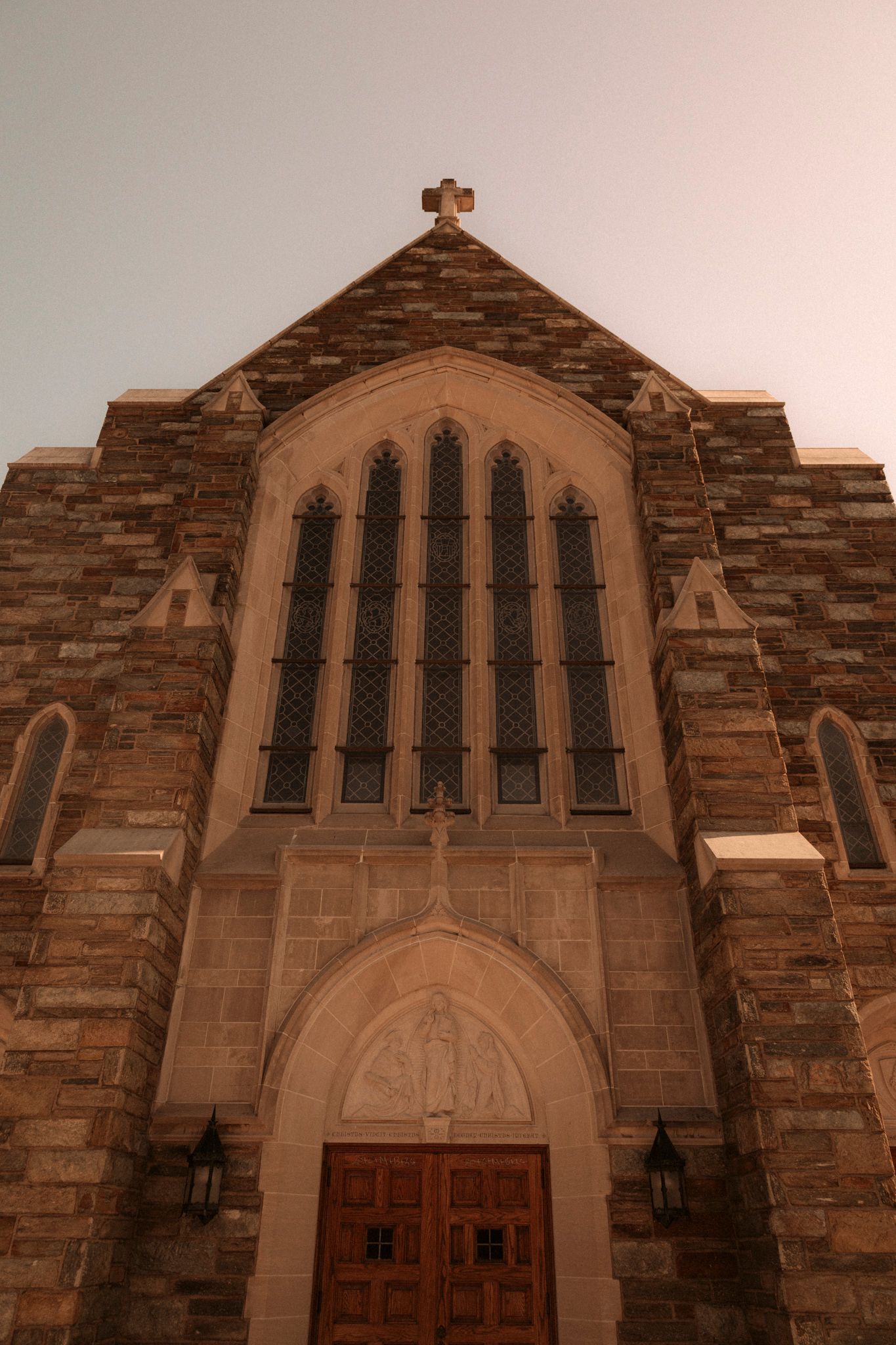 Gothic stone church facade with arched stained glass windows, wooden doors, and a cross atop the peaked roof.