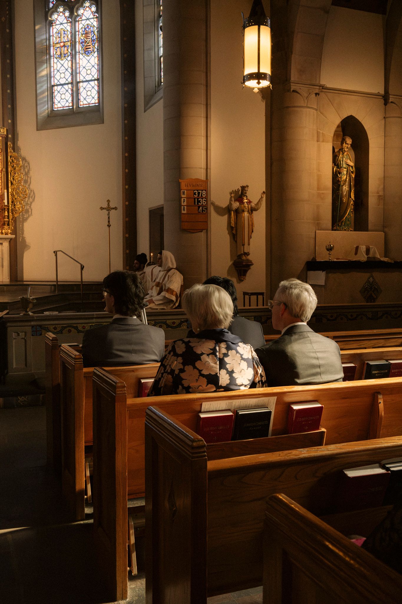 Elderly wedding guests seated in wooden church pews during a ceremony, with stained glass windows and altar visible.
