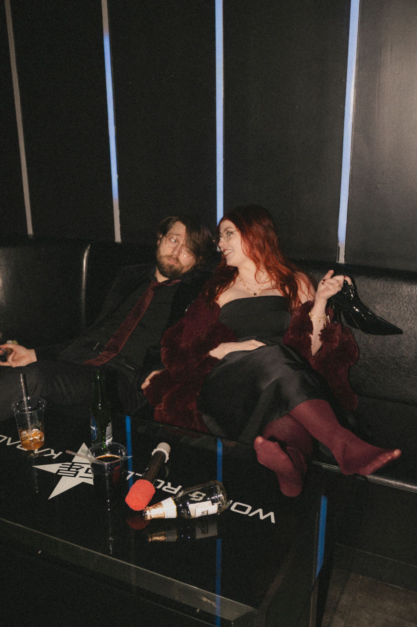 Two people relax on a dark leather couch at what appears to be a lounge or club setting with dim lighting.
