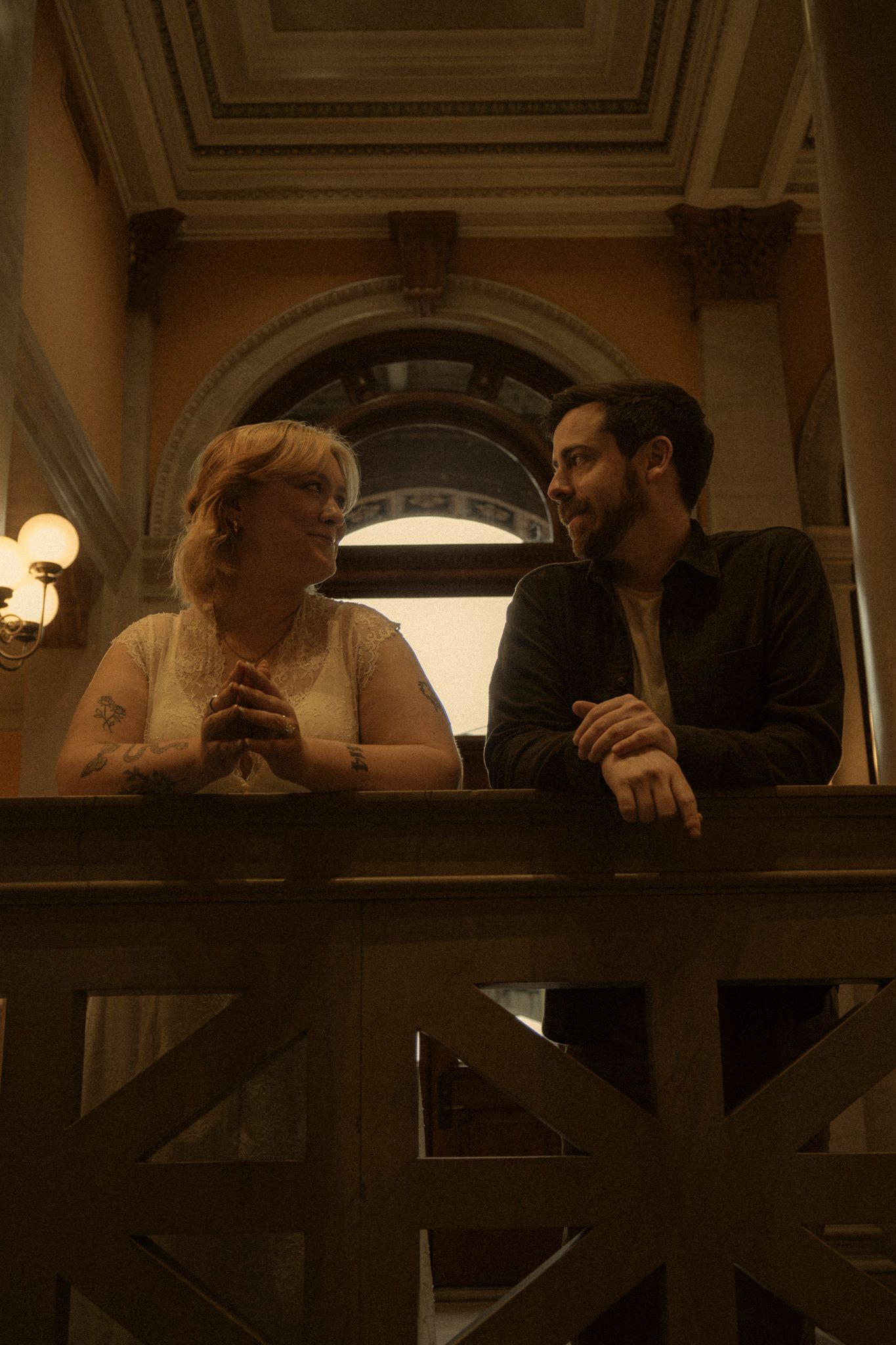 Two people lean over a railing in a dimly lit interior space with ornate architectural details.