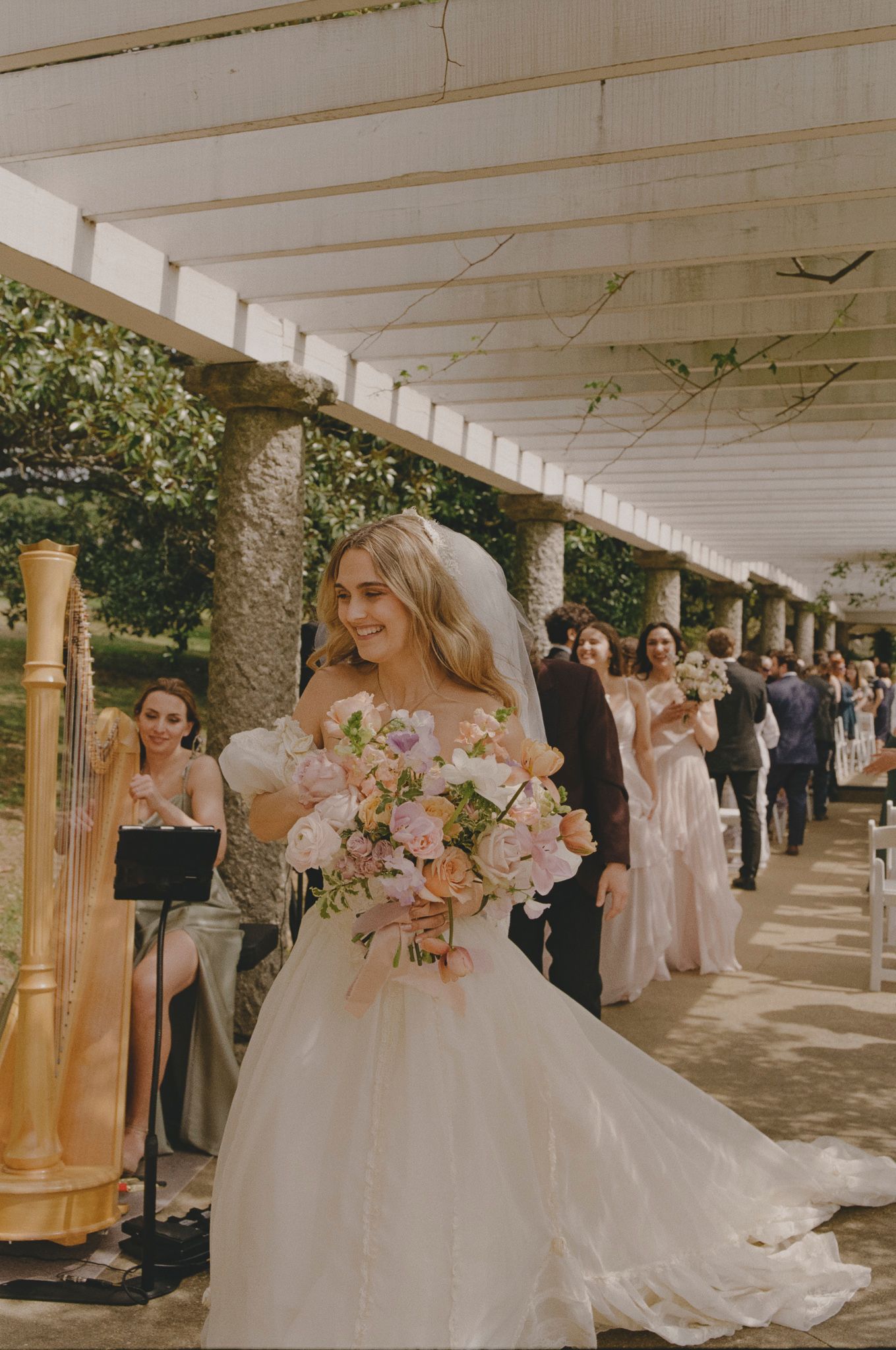 Wedding party gathered under a white pergola walkway with stone columns during an outdoor ceremony.