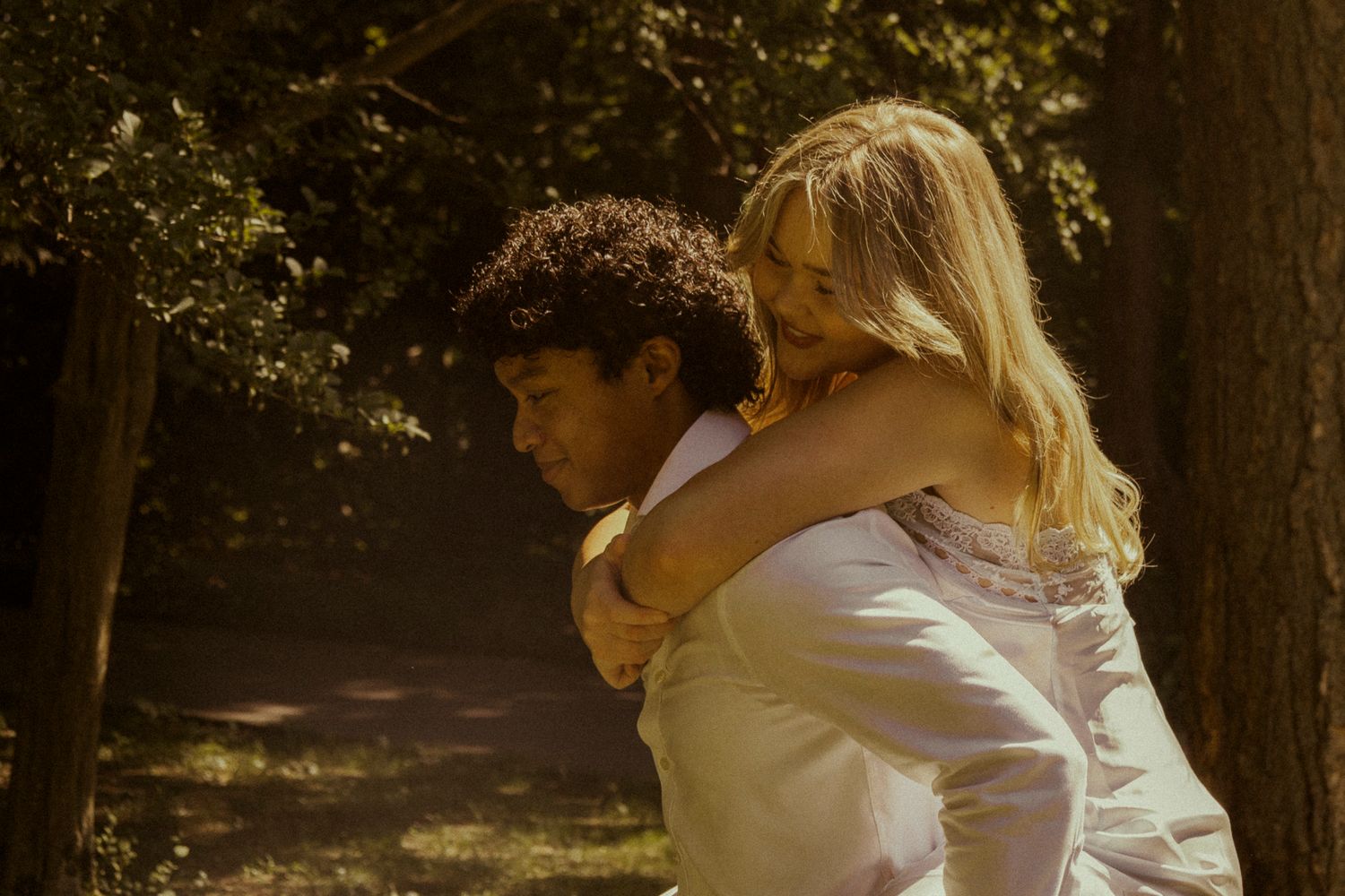 A groom gives his laughing blonde bride a piggyback ride in a sun-dappled forest during their wedding day.