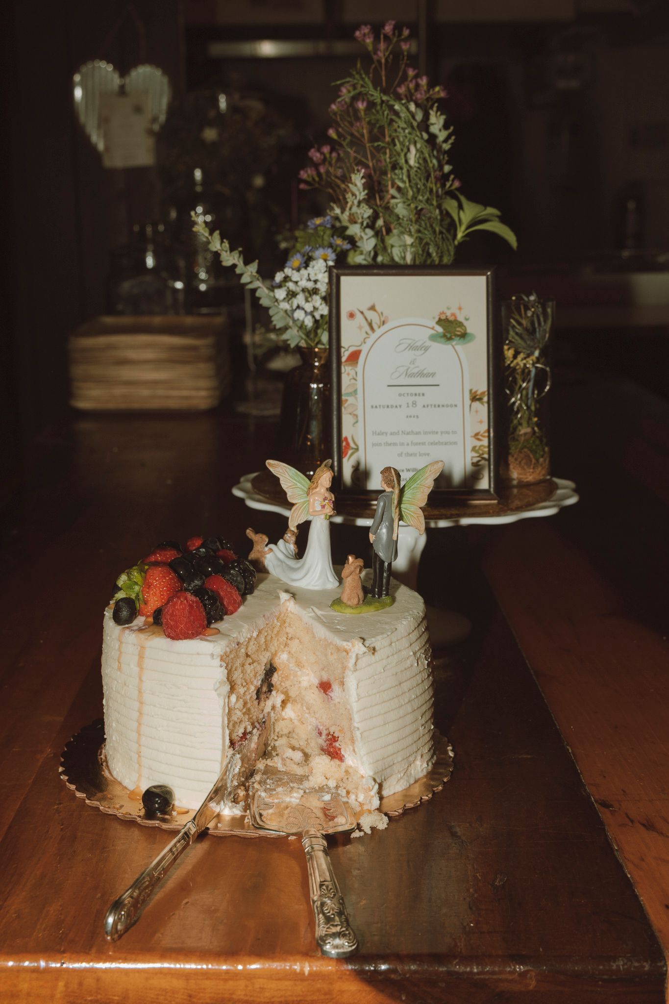 A rustic wedding cake topped with strawberries and a decorative bird figurine sits beside floral arrangements on a wooden table.