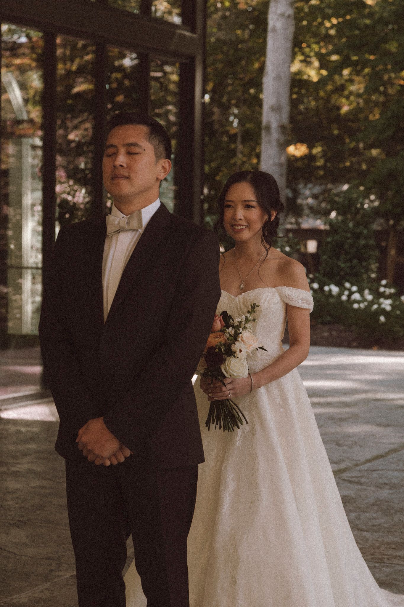 A bride in an off-shoulder white gown and groom in black suit share a tender moment in a wooded outdoor setting.