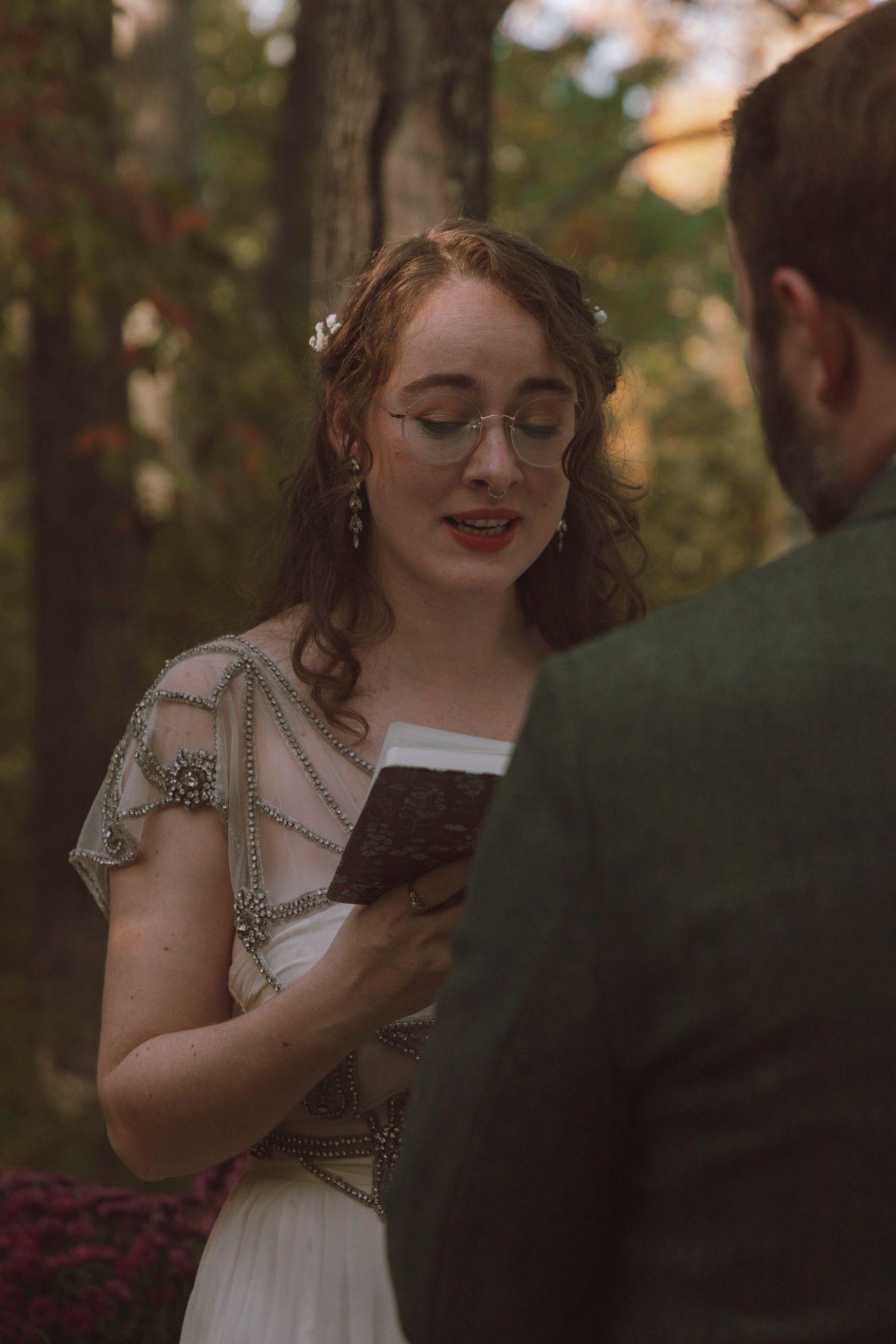 A person in a vintage-style dress smiles while holding a chocolate bar in a wooded outdoor setting.