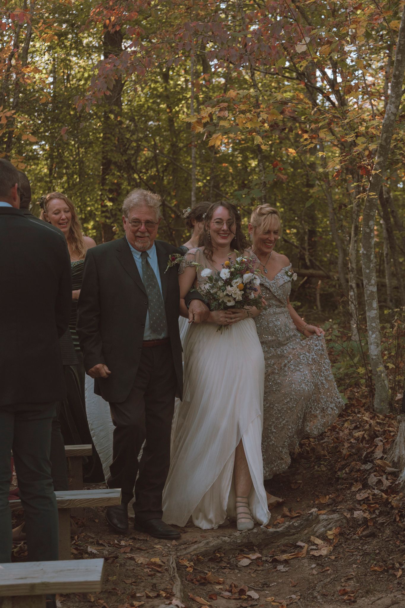 A wedding ceremony takes place on a wooden walkway in an autumn forest with warm golden light filtering through trees.