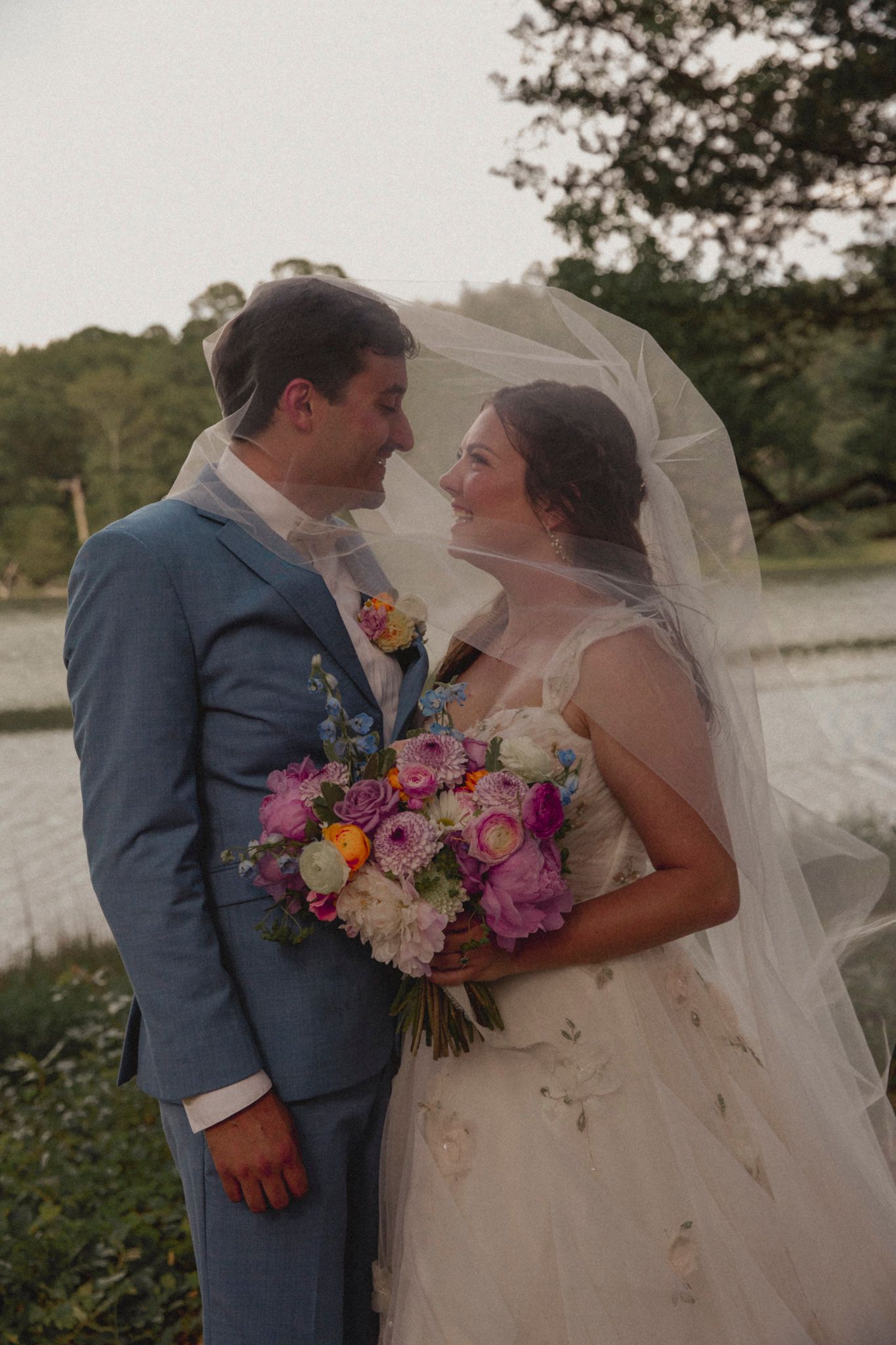 A romantic wedding couple embraces outdoors with the bride in white holding a vibrant pink and orange bouquet.