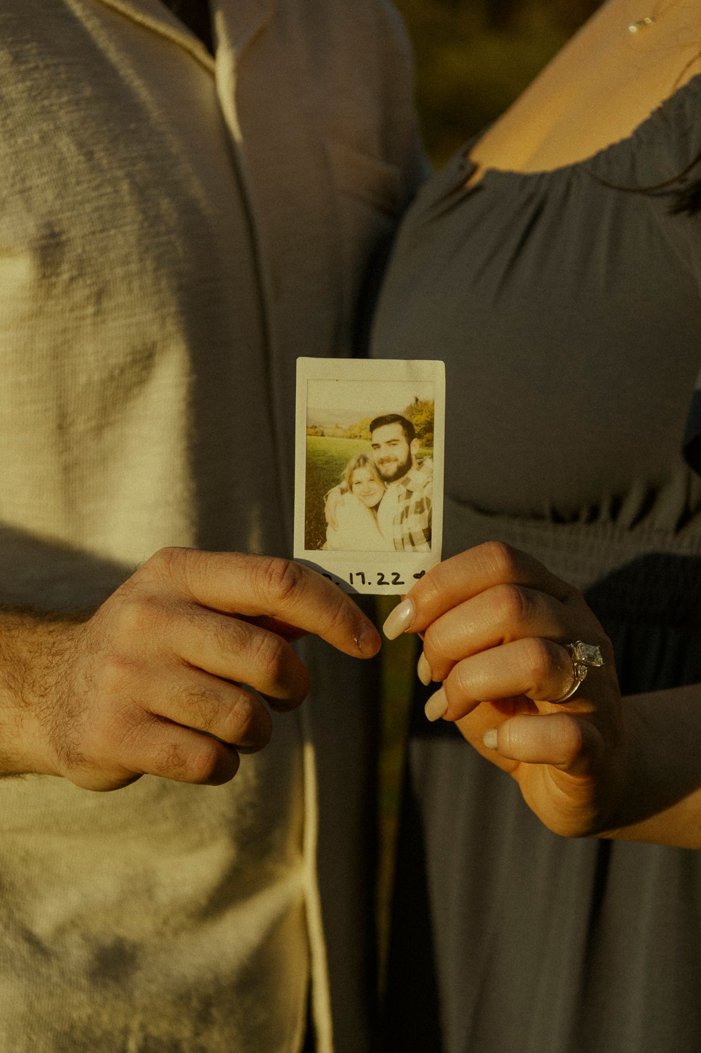 Couple holding a Polaroid photo together outdoors, woman wearing an engagement ring at golden hour.