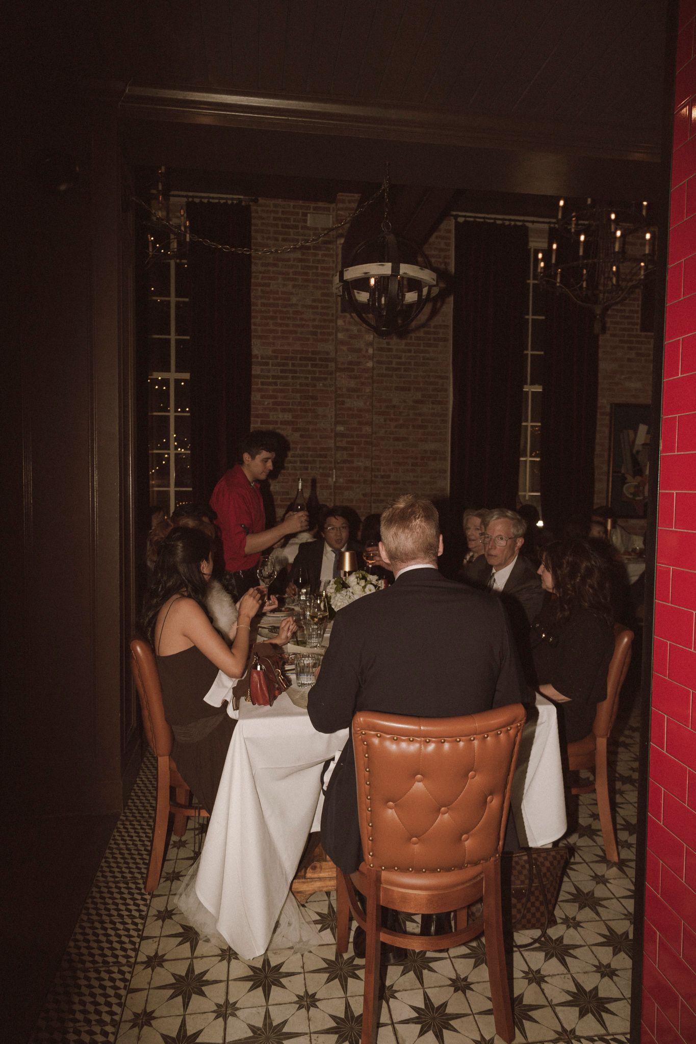 Wedding guests gathered around a candlelit dinner table in a warmly lit restaurant with brick walls and patterned floors.