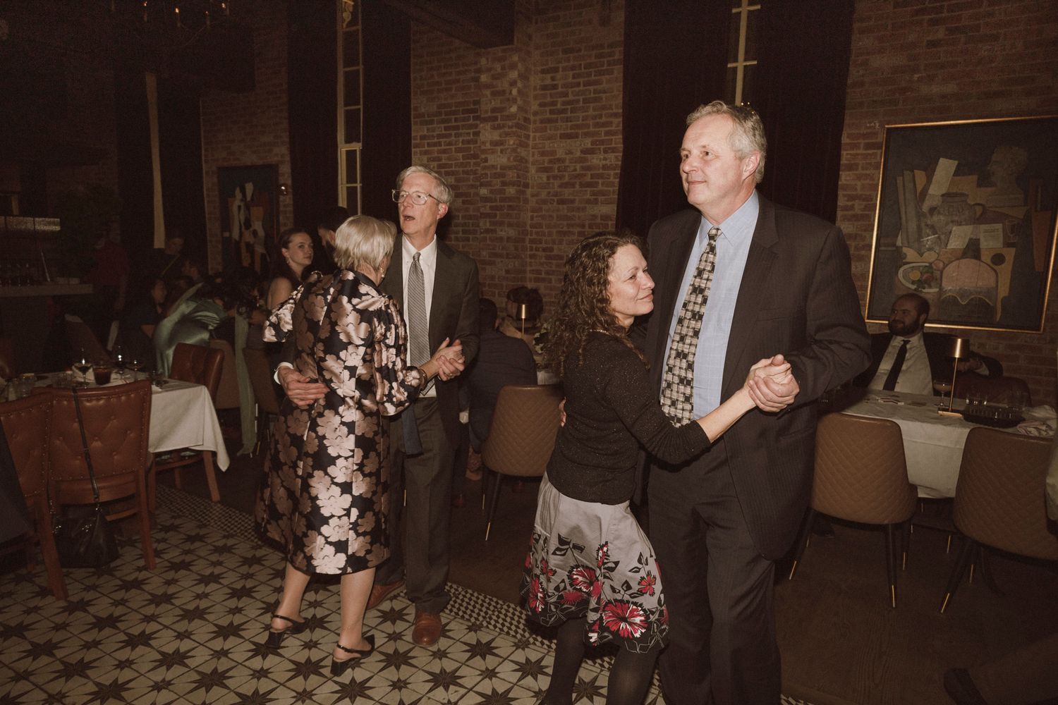 Two couples dancing together at wedding reception in dimly lit brick-walled ballroom.