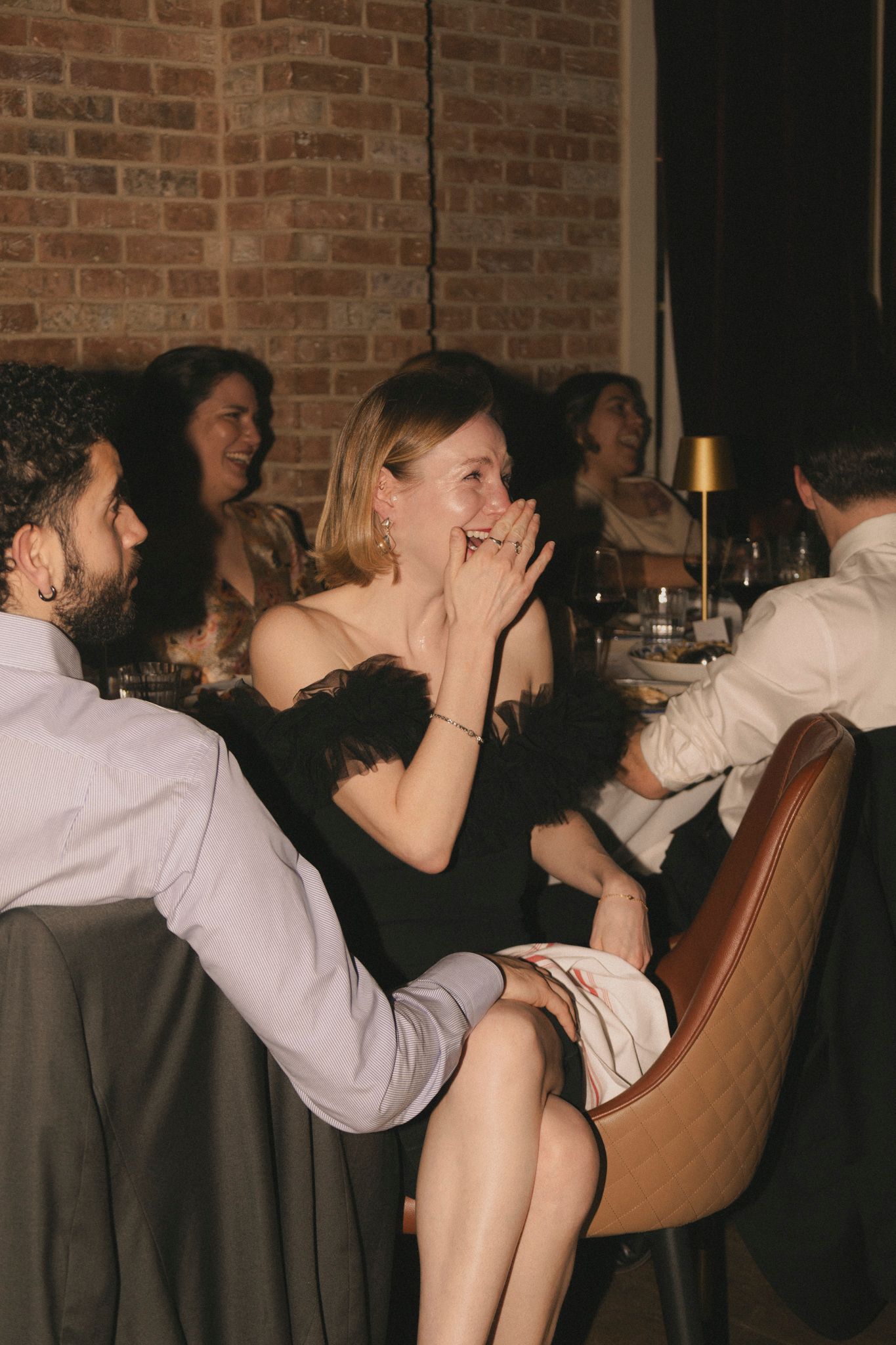 Woman in black feathered dress laughing joyfully with guests at a dimly lit wedding reception dinner table.