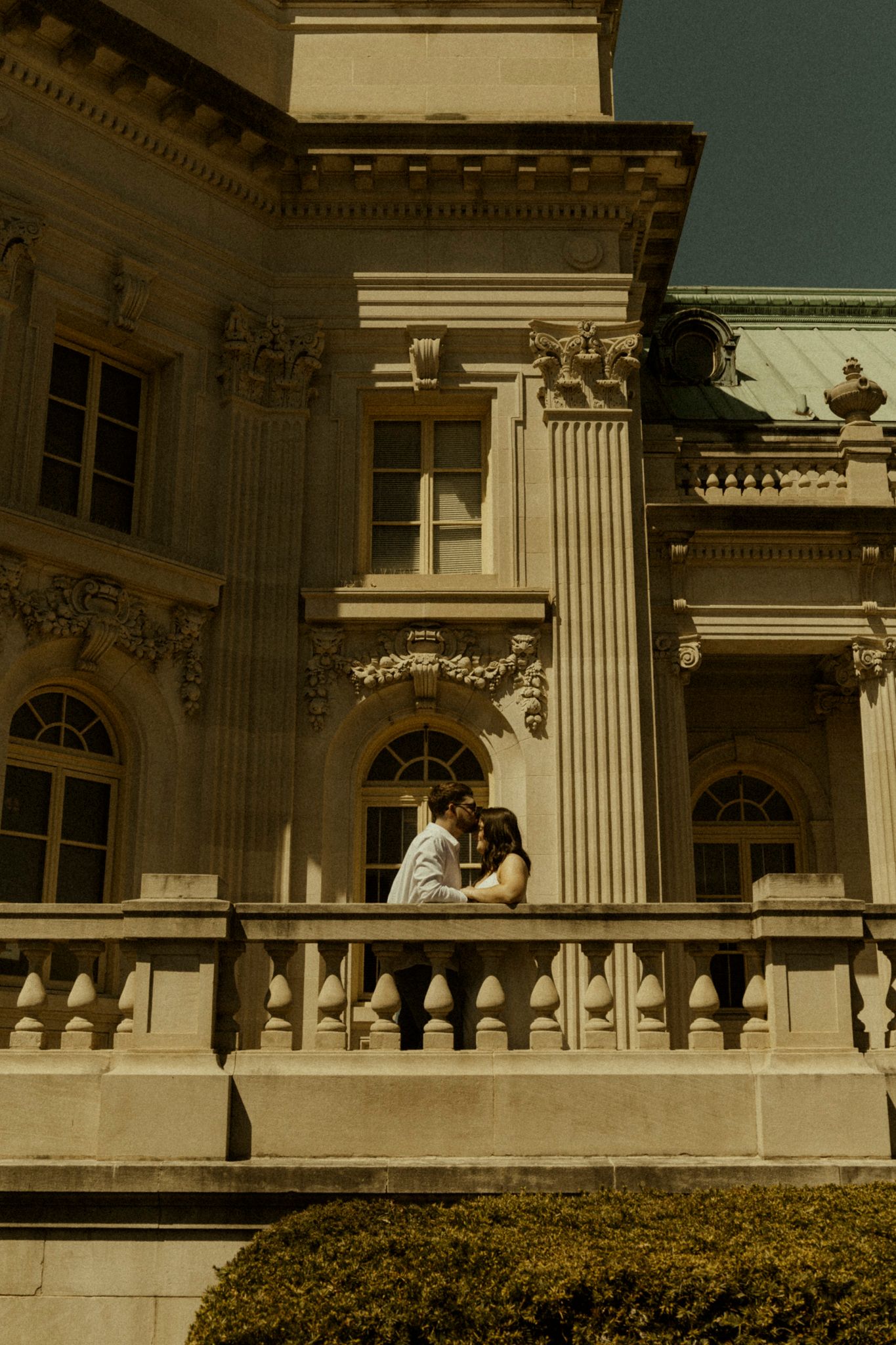Couple embracing on ornate stone balcony of elegant neoclassical building with decorative columns and arched windows.
