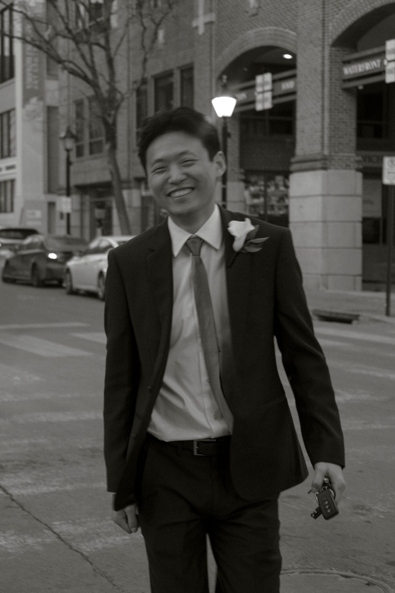 Smiling groom in a dark suit with boutonniere walking on a city street in black and white photo.