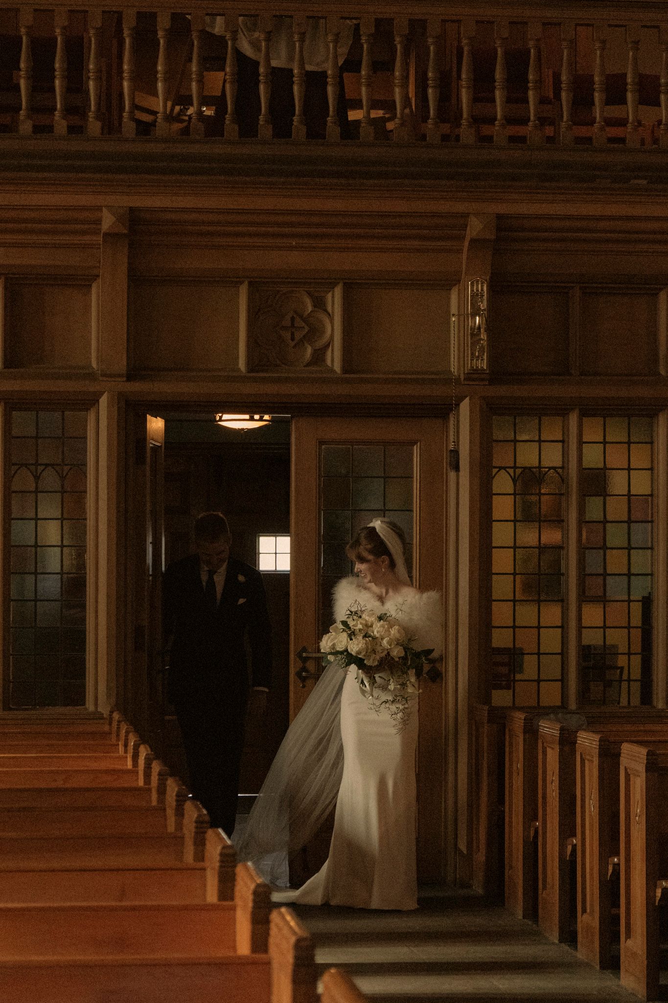 Bride in white gown and veil holding bouquet enters church doors with escort during wedding ceremony.