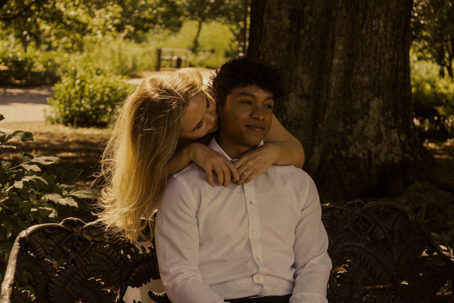 A couple sits under a tree, woman with blonde hair resting her head on a man's shoulder in a sunny park.