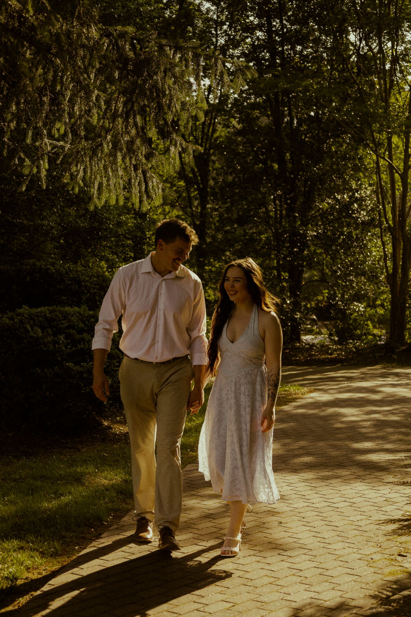 Couple holding hands walking on brick path through sunlit green forest.