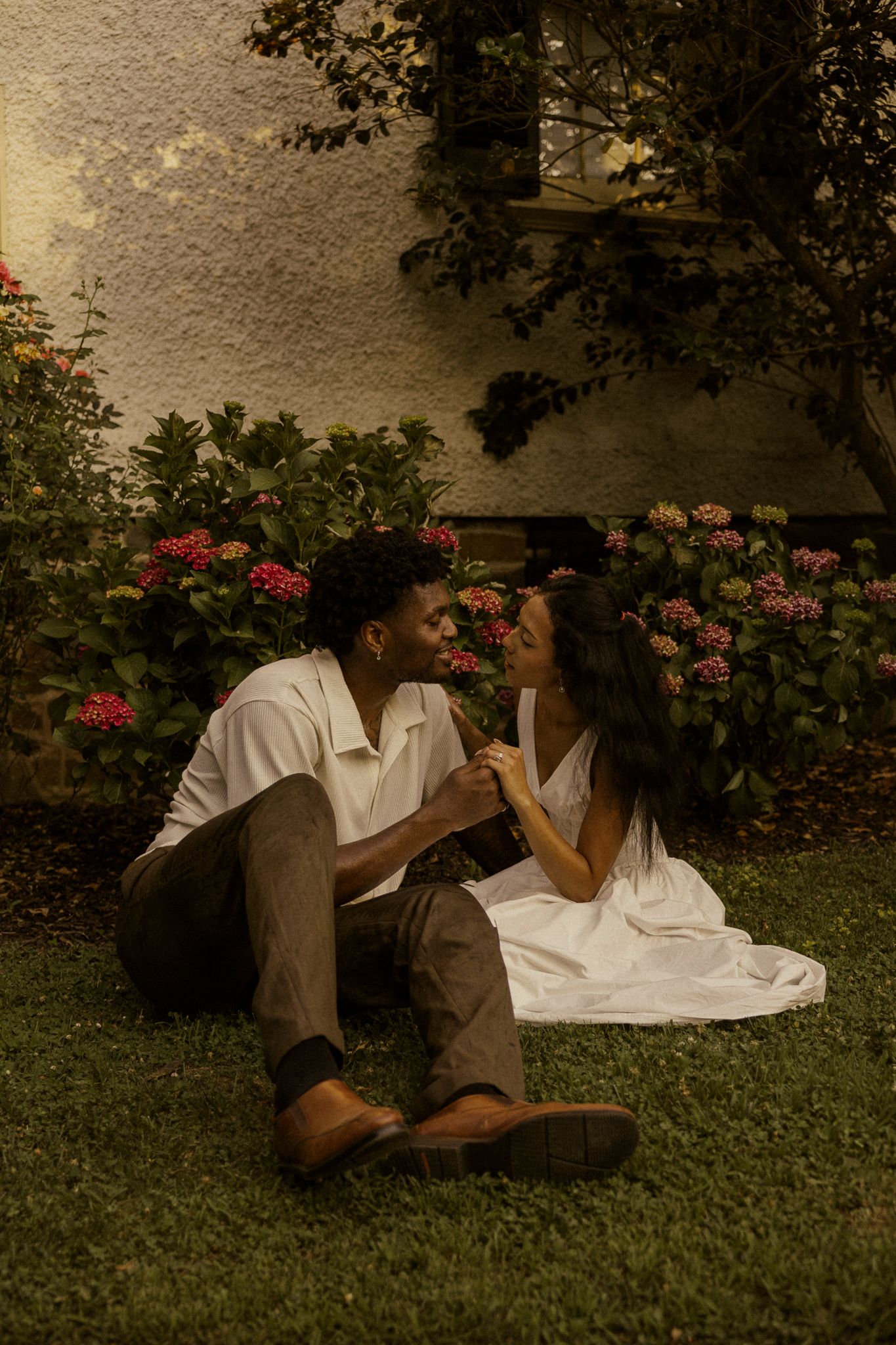 A couple sits on green grass in a garden, holding hands and gazing at each other near colorful flowers.