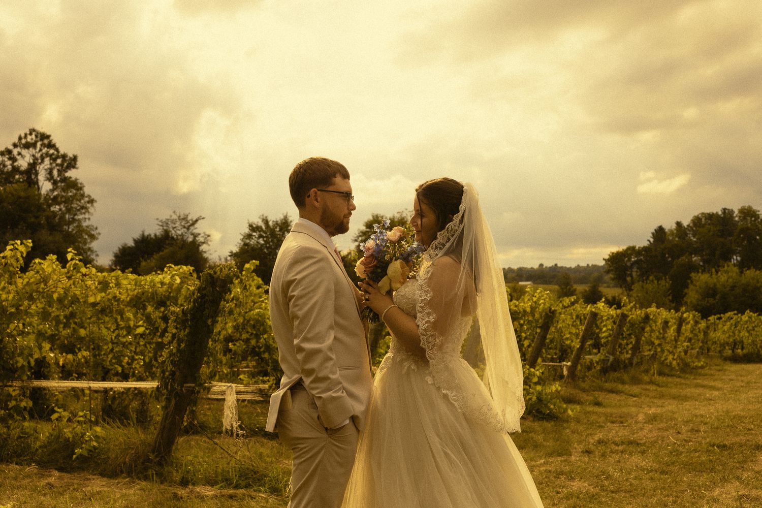 Bride and groom share a romantic moment in a vineyard, with lush green vines and dramatic cloudy sky backdrop.