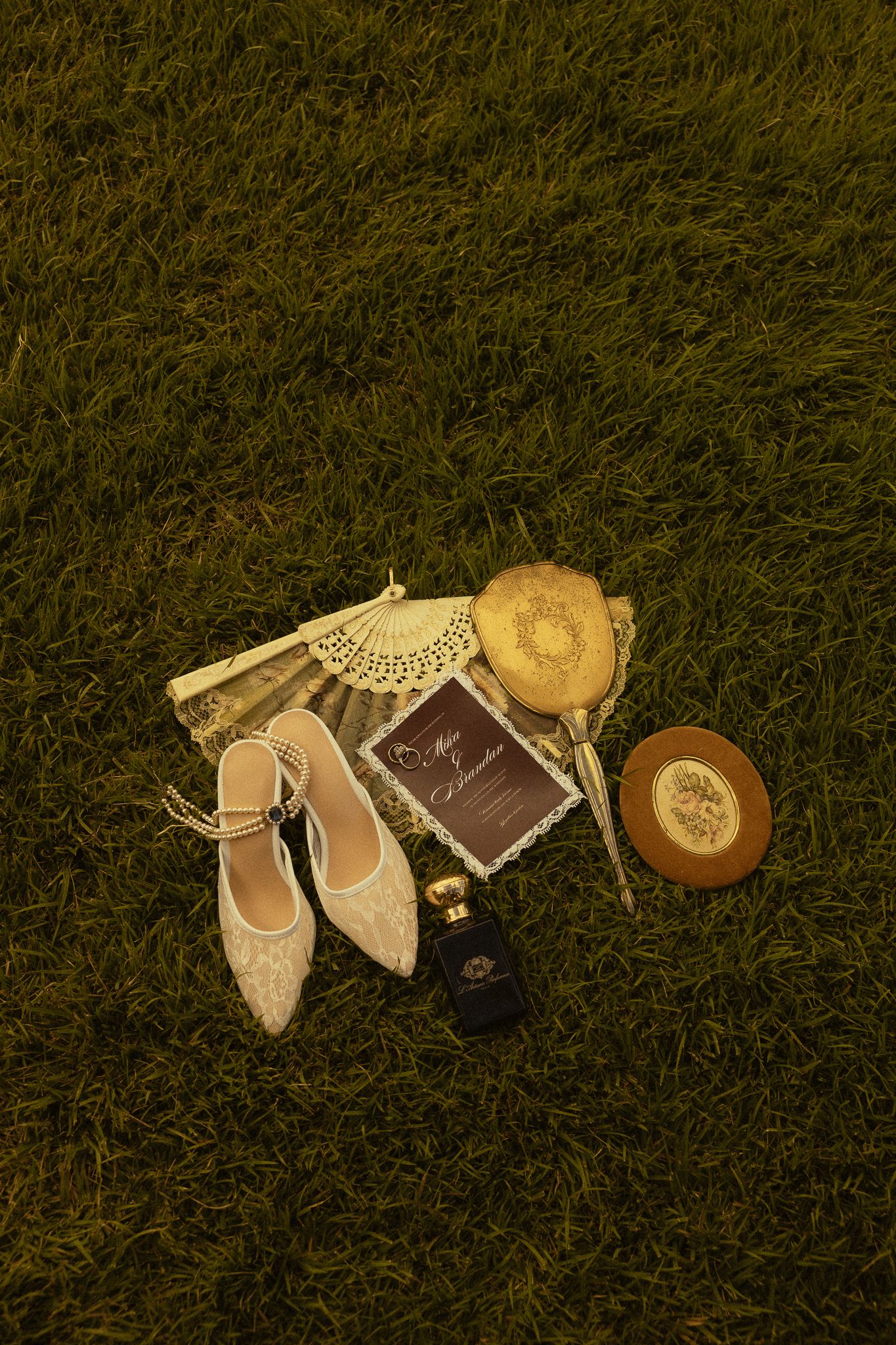Elegant bridal accessories on grass, including lace flats, perfume, gold purse, fan, and wedding invitation.