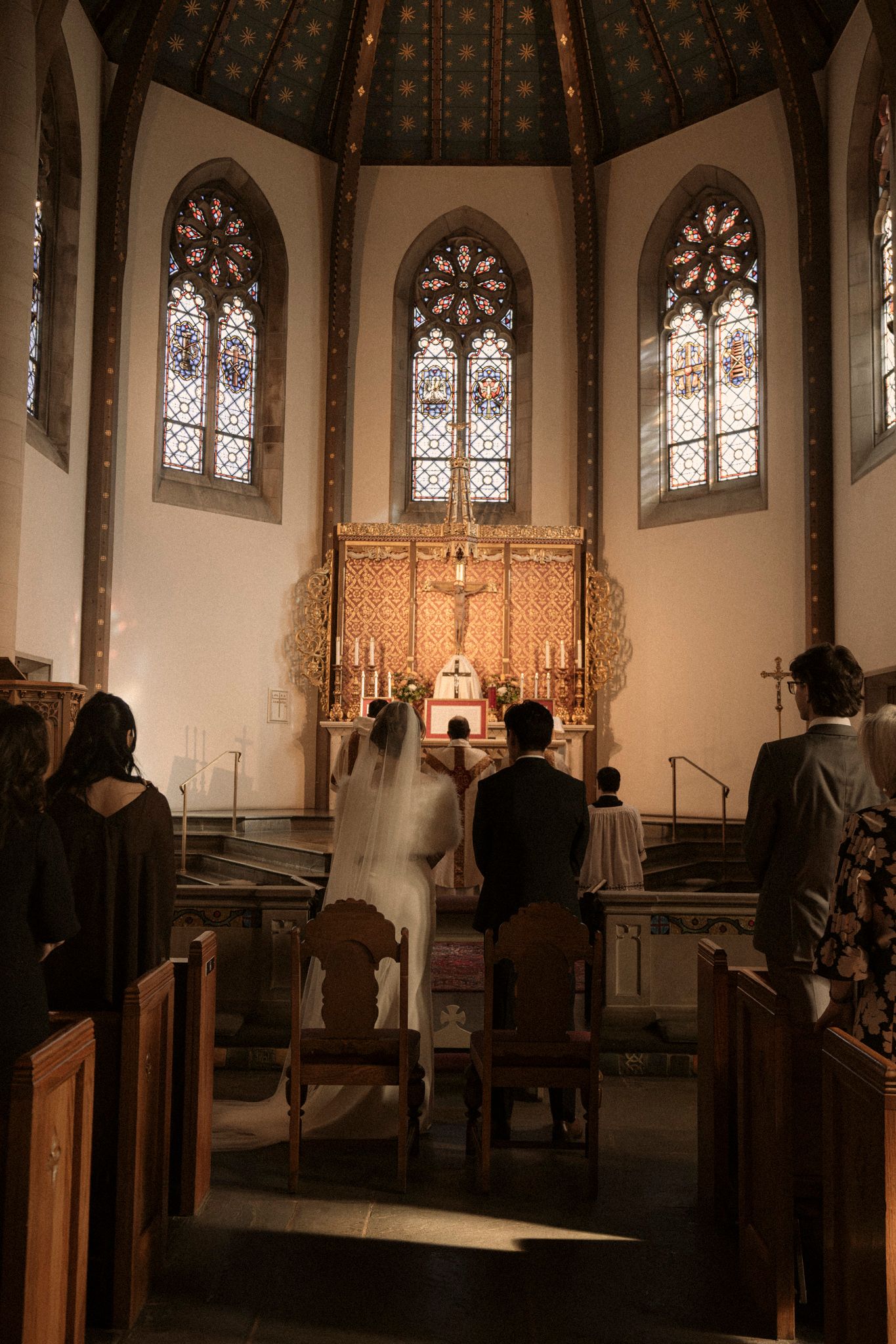 A traditional church wedding ceremony taking place in a Gothic chapel with stained glass windows and an ornate golden altar.