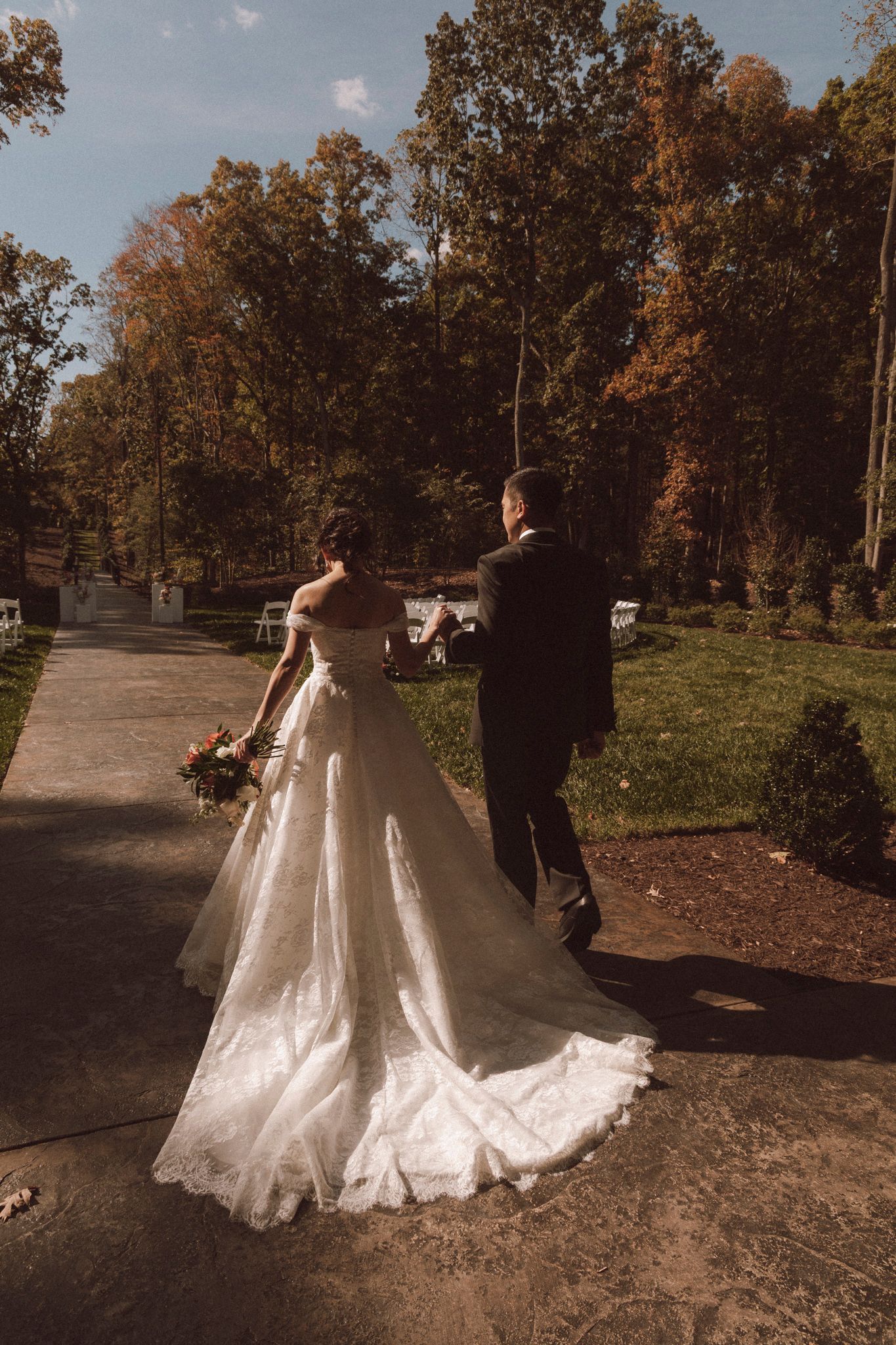 A couple in wedding attire walking down a tree-lined path during autumn with a trailing white wedding dress.