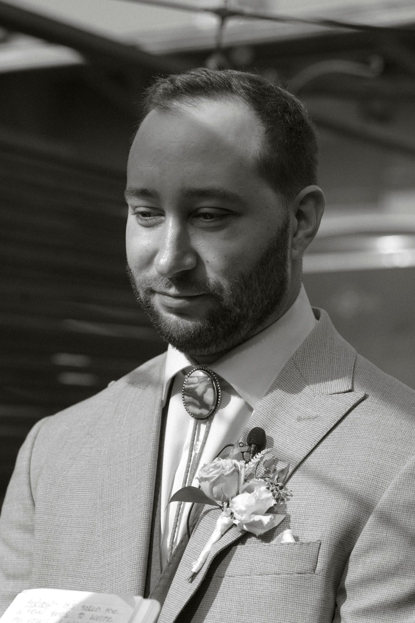 A groom in a beige suit with floral boutonniere smiles during wedding ceremony.