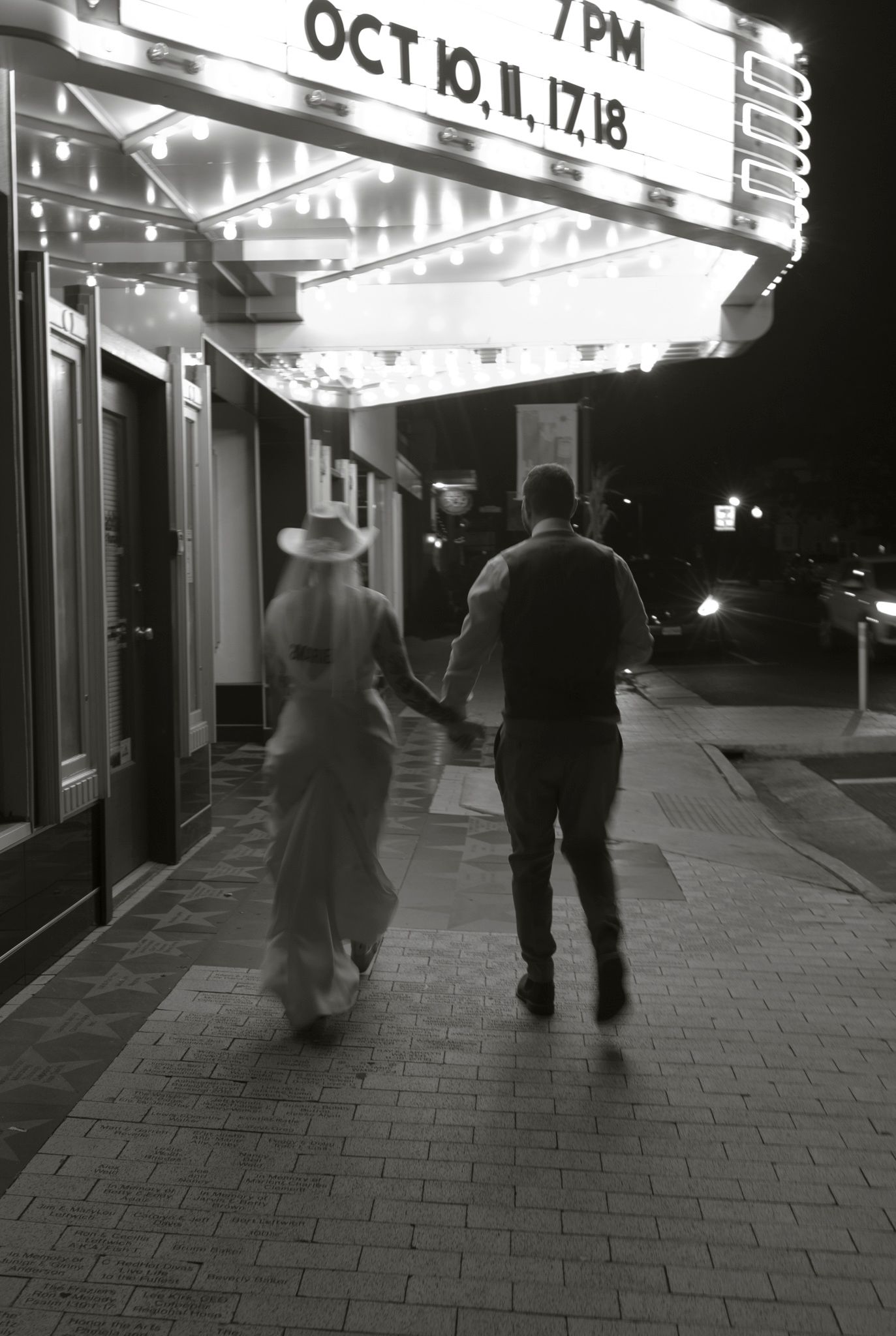 People walking on a dimly lit sidewalk at night with illuminated storefronts and marquee lights visible in the background.